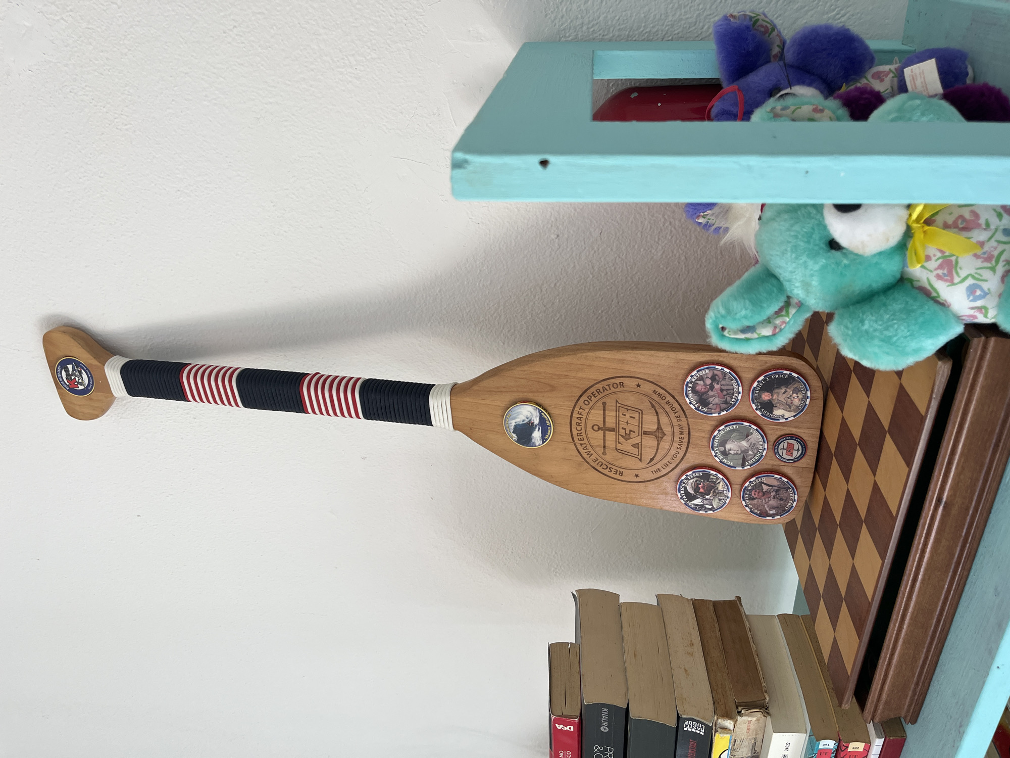 The image shows a decorative paddle mounted on a white wall within a room. The paddle is primarily light brown wood, with a dark blue and red striped grip on the handle. There are several circular pins attached to the blade of the paddle. These pins appear to have images of people, possibly military or service personnel. The paddle is inscribed with text, including "RESCUE WATERCRAFT OPERATOR." Below the paddle, there are several books stacked on a shelf. Above the paddle, there's a blue shelf containing some stuffed toys including a purple plush toy and a mint-green teddy bear. There's a brown chess/checkerboard. The location in Santeru/San Vero Milis, Italy is not ascertainable from the image.