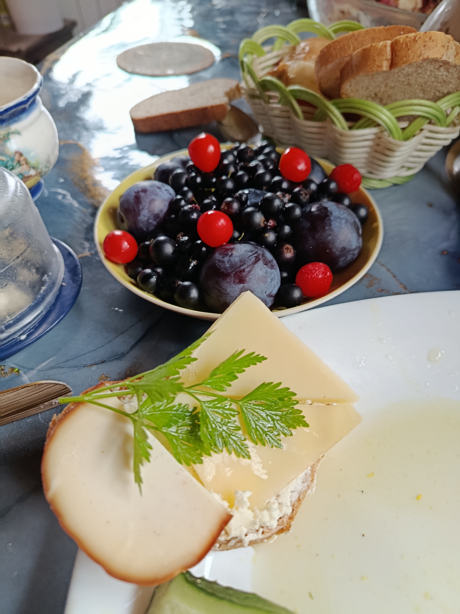 The image depicts a still life of food items arranged on a table, likely in a home setting in the city of Vereya, Russia. In the foreground, a slice of bread is topped with a spread, a slice of cheese, and a sprig of parsley. To the right, a white plate contains a creamy liquid, possibly soup or a sauce, with a sliver of cucumber visible at the bottom. In the midground, a yellow bowl is filled with black currants, plums, and small red berries, possibly cranberries. Behind this, a woven basket holds slices of bread. A decorative pitcher and a glass cloche are also visible. The table surface appears to be a dark, textured material, possibly a tablecloth, with reflections on its surface. The lighting suggests an indoor environment, potentially during the day. There are no people visible in the image.