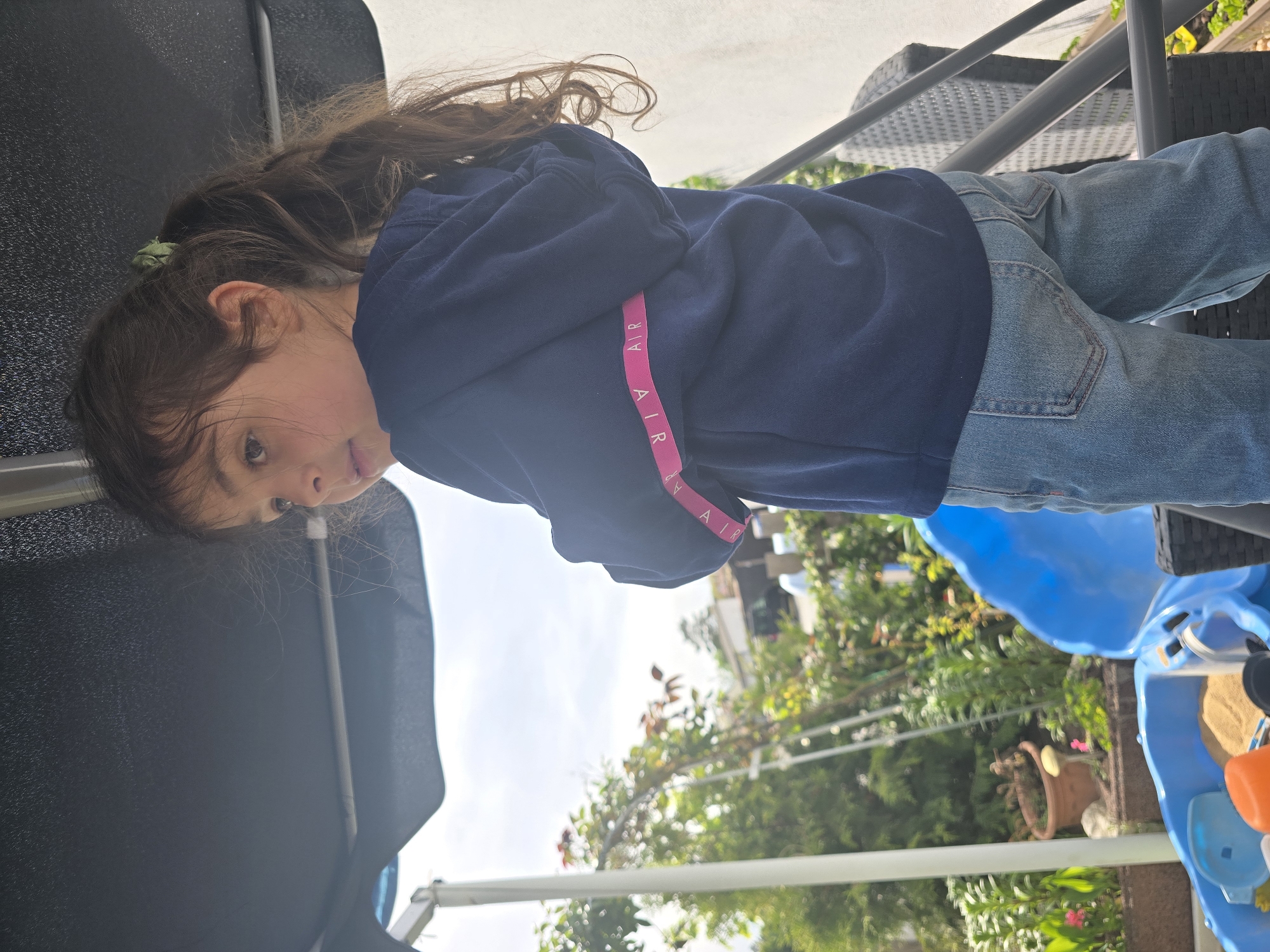 A young girl with long brown hair sits wearing a blue hoodie with pink AIR branding and blue jeans. The background includes green foliage and a blue sky, suggesting an outdoor setting, likely in a backyard. There's a blue water table visible, along with patio furniture and other garden features. Given that the instructions ask that it be located in Zirndorf, Germany, the image is most likely taken there.