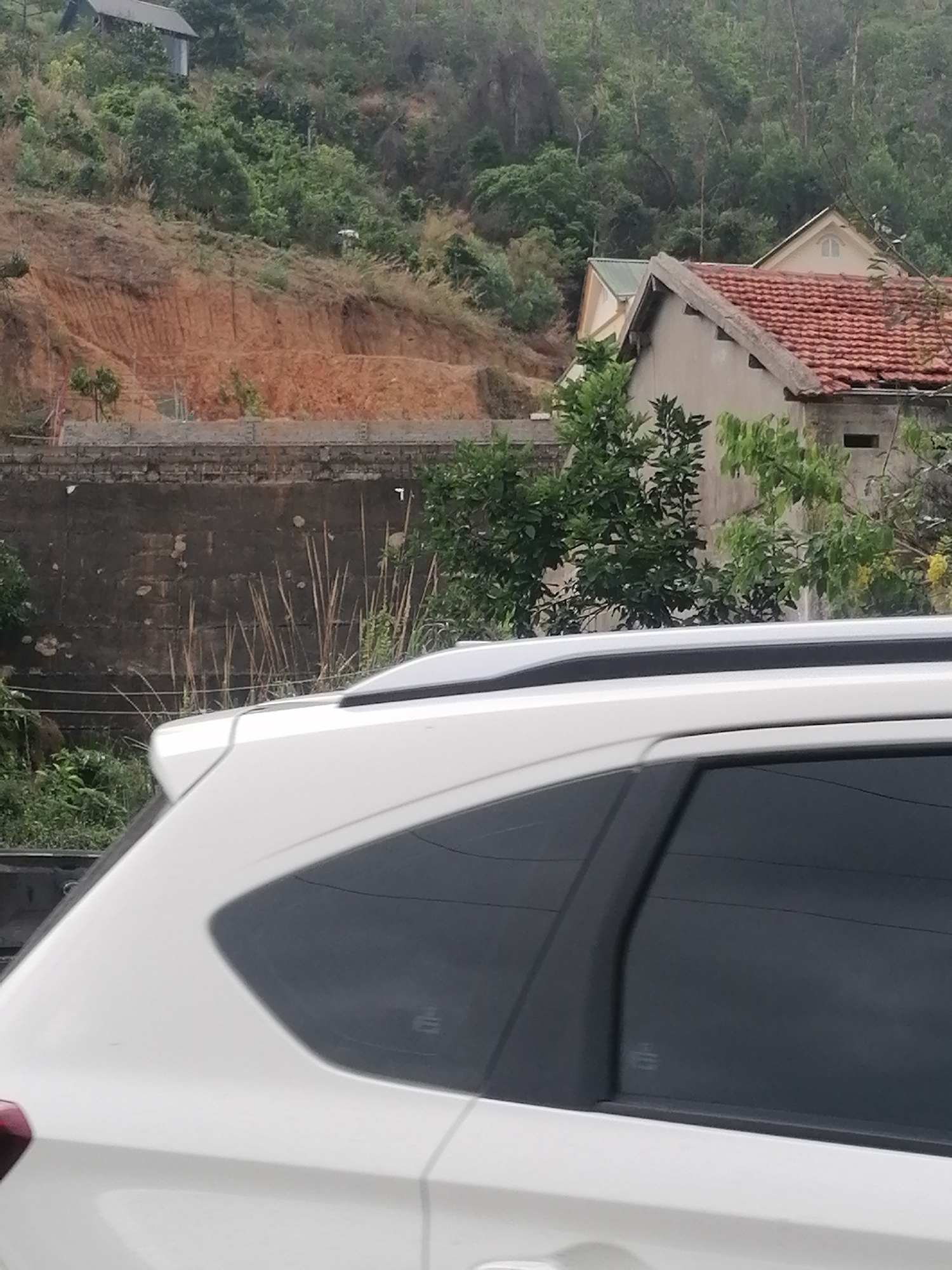 The image presents a daytime scene dominated by a white car in the foreground and a hilly landscape with buildings in the background. The upper rear section of a white SUV or crossover is visible, featuring black roof rails and dark-tinted rear side windows.
A dark, weathered retaining wall or concrete barrier runs horizontally across the middle ground, with tall, dry grasses and other sparse vegetation growing directly in front of it. Beyond this wall, a steep hillside rises, showing significant patches of exposed reddish-brown soil, suggesting erosion or recent earthworks. The hillside is also covered with a mix of green trees and drier vegetation.
In the background, nestled on the hillside, are several structures. A small, rustic building with a dark roof can be seen high up, partially camouflaged by trees. Closer to the mid-ground, there are houses featuring traditional terracotta-tiled roofs; one house prominently displays a light-colored gable end with an apparent attic window.
The lighting is diffuse, indicating an overcast or hazy day, as there are no distinct shadows. The scene depicts a rural or semi-urban environment in a hilly region, aligning with the provided location context of Bai Chay Ward, Vietnam, known for its mountainous terrain. No people or visible text are present in the image.