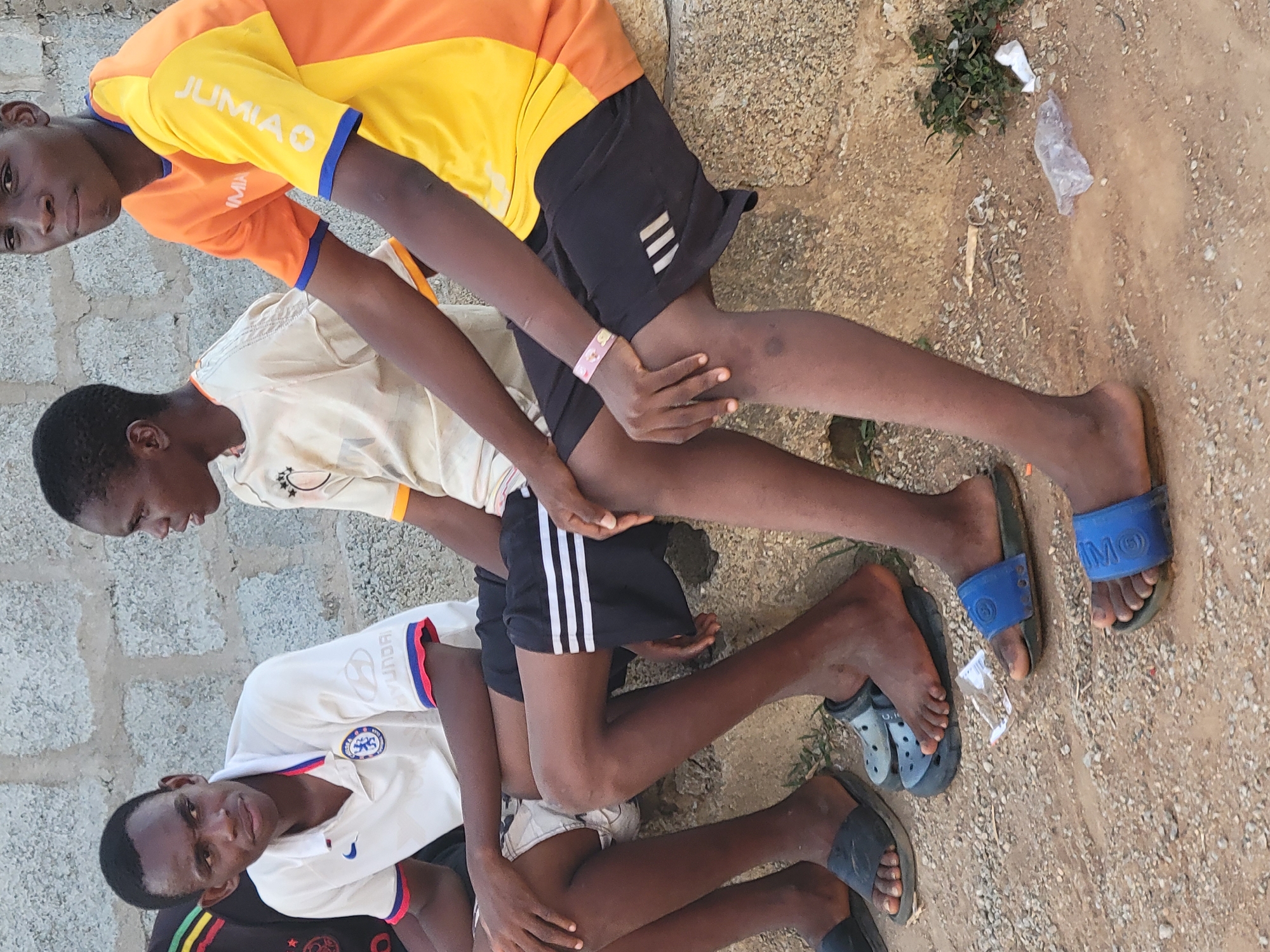 Three young men are sitting or kneeling in a row on a dirt surface, possibly in Ibo, Nigeria. The man in the foreground wears a white Chelsea football shirt with red and blue trim, sitting behind the other two. The middle man is wearing a tan shirt and black shorts. The top man wears a yellow and orange shirt with the word "JUMIAO" on the sleeve, and black shorts with white stripes. The dirt ground is scattered with small stones and debris, including a plastic bag. The composition is slightly rotated.