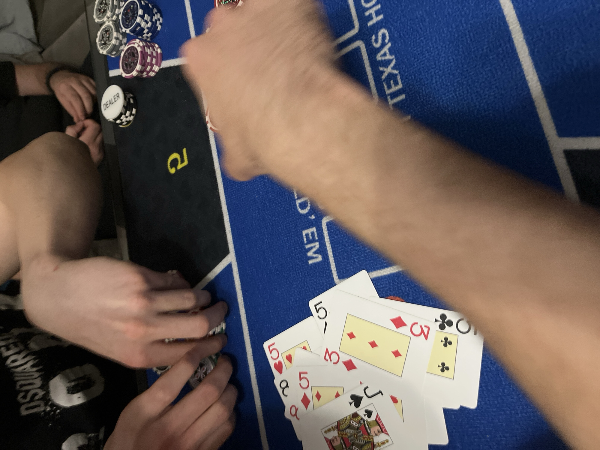 The image depicts a casual game of Texas Hold'em poker underway in an indoor setting in Burgas, Bulgaria. At least three individuals are visible, though mostly through their hands, arms, and legs, suggesting an intimate and focused gathering.
The game is played on a blue poker mat, which distinctly displays the text "TEXAS HOLD'EM". Various stacks of poker chips, including white/black, blue/white, and purple/white combinations, are neatly arranged on the left side of the mat. A prominent white chip marked "DEALER" rests atop a stack of black and white chips. In the foreground on the right, several playing cards are fanned out, revealing some of their values: multiple fives (5 of hearts, 5 of diamonds, 5 of clubs), a 3 of diamonds, a Queen of clubs, and a Jack of spades. One person's arm extends from the right, appearing to interact with the cards, while another person's hands are seen near the chips on the lower left. A third participant's leg and arm are visible on the far left.
The activity is clearly a poker game, with players engaged around the table. The atmosphere seems relaxed, typical of a home game. One person's black t-shirt shows partially visible text "DSQUARED". The lighting indicates an indoor setting, likely evening or night. No specific weather details are discernible from this indoor shot.