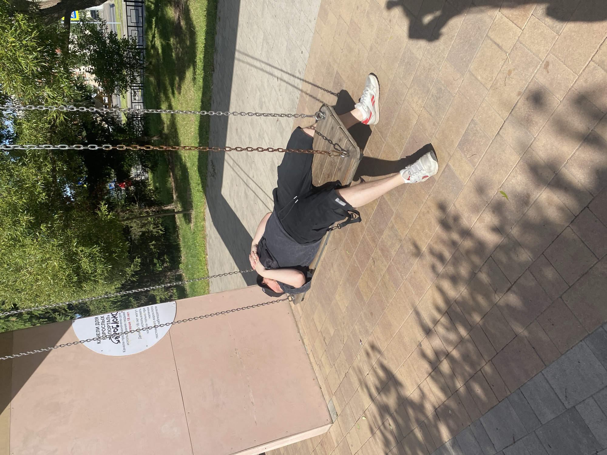 A person lies on a wooden swing in a public park, their head resting on their hands. They are wearing a dark t-shirt, black shorts, and white athletic shoes with red accents. The swing is suspended by metal chains attached to an unseen support structure. Behind the swing, a light pink, slanted panel leans against a wall, with a circular white sign affixed to it. The sign contains text in Russian, which appears to be instructions or information related to the swing. The ground is paved with interlocking tan and gray bricks, with shadows of trees cast upon them. In the background, lush green trees and foliage are visible, along with a street and a building. The scene suggests a sunny day in an outdoor recreational area. The sign can be translated to read "SWINGS FOR ADULTS" and "PORTAL GOROBOBROTSK". It further states that the swings are intended for adults, and children under 18 can use them only with adult supervision.