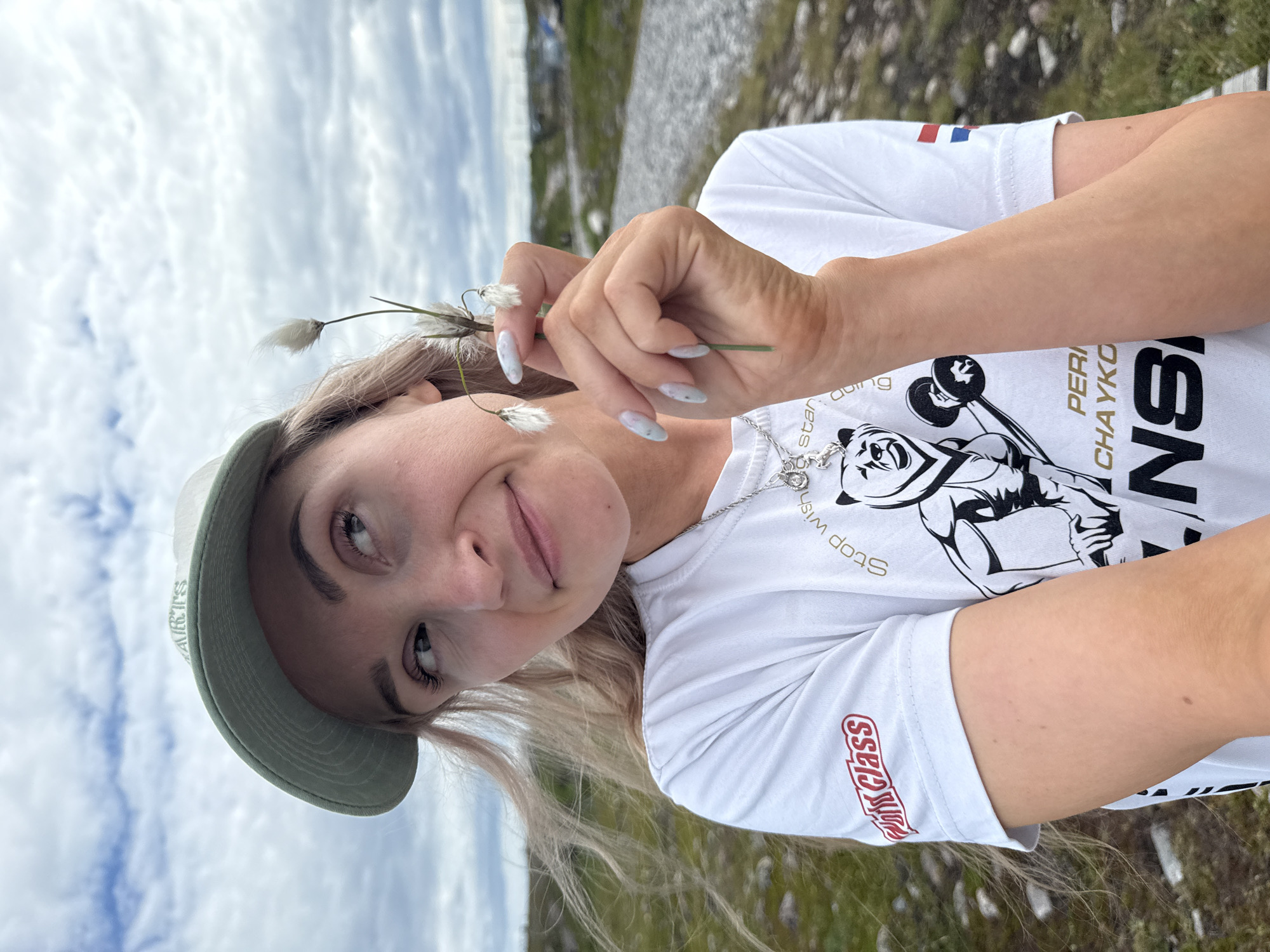A young woman with blonde hair and light skin wears a white t-shirt with graphics of a bear lifting weights and text. She is also wearing a light green baseball cap. She is holding a delicate white plant with fluffy seed heads in her right hand, which is positioned near her face. Her fingernails are light blue with a speckled pattern. The background appears to be a natural landscape with a cloudy sky, grass, rocks, and a gravel path, consistent with the environment of Teriberka, Russia. The overall scene captures a casual, outdoor moment with the woman interacting with nature.
