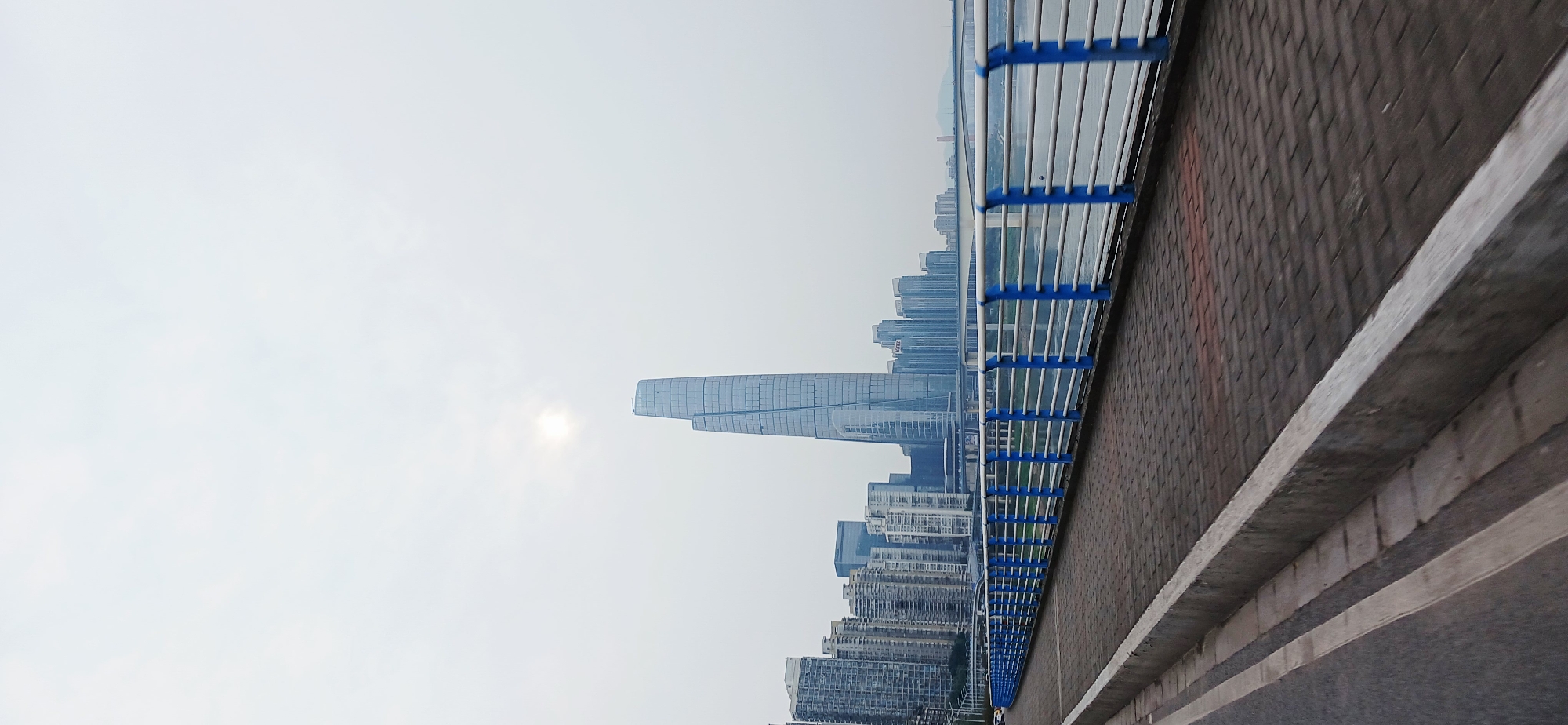 The image depicts a view of a cityscape in Yuzhong District, Chongqing, China, from an elevated position, potentially a bridge or overpass. Predominantly, the scene showcases modern high-rise buildings clustered together against an overcast sky. One distinctively tall skyscraper stands out with a stepped design near the top. Along the lower right side of the image runs a concrete structure with a guardrail made of blue and white steel, indicating the vantage point. The foreground features a road surface. No people are visible in the scene.
