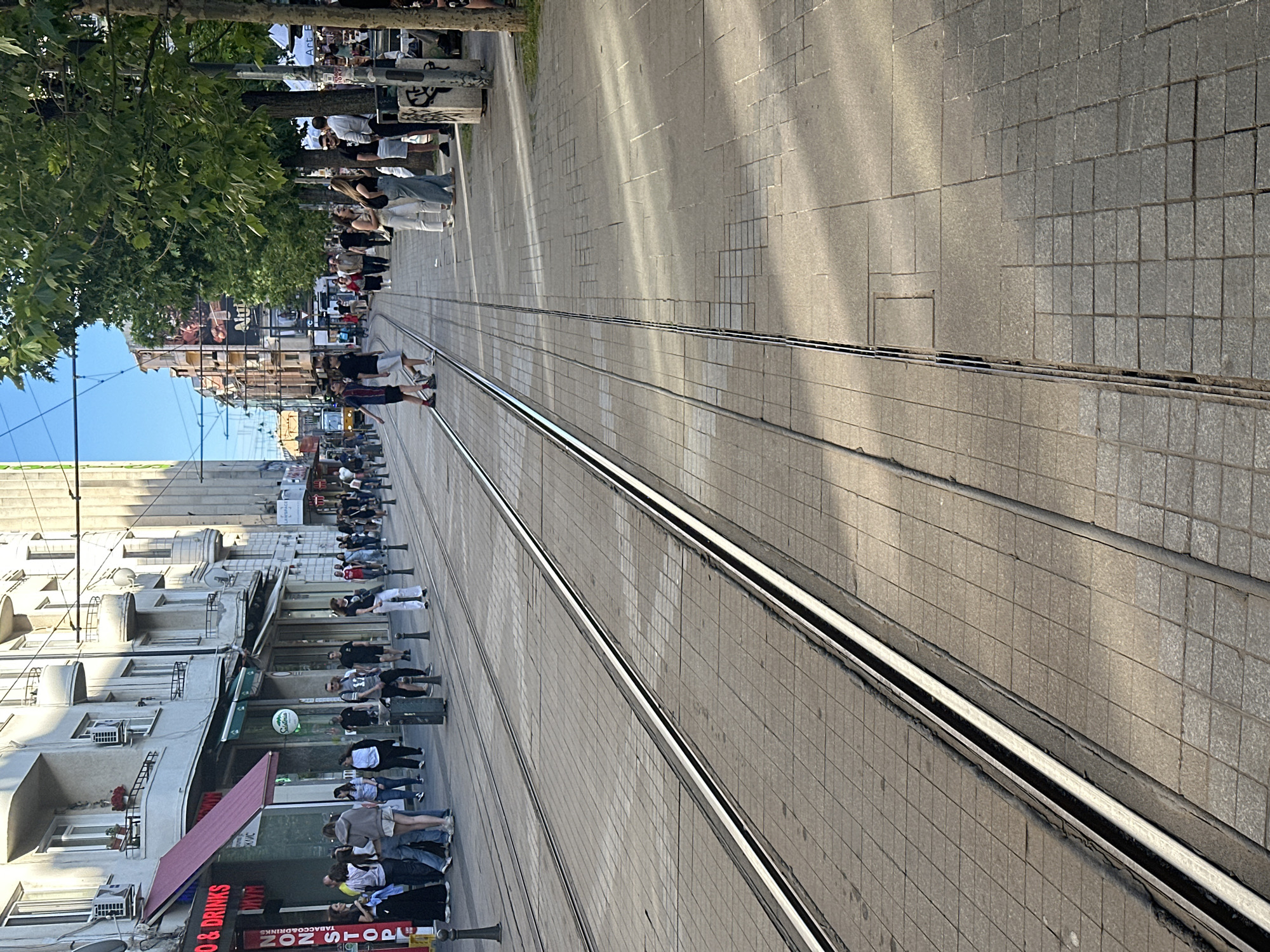 A street in Sofia, Bulgaria, featuring tram tracks running through a pedestrian area. People are walking along the street and are standing by businesses and trees. Buildings line the street. The image is taken from ground level alongside the tram tracks, looking down the street.