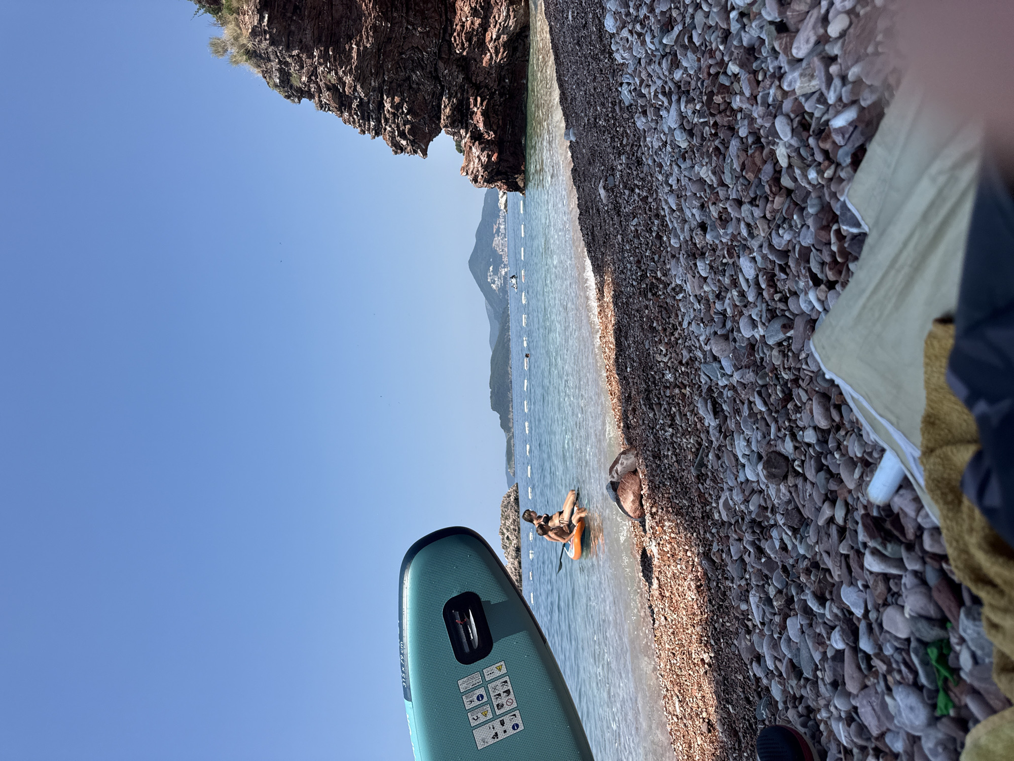 The image depicts a sunny day at a pebbled beach in Boreti, Montenegro. In the foreground, a teal stand-up paddleboard rests on the shore, its brand and size markings visible: "315 x 79 x 15 cm". The paddleboard also displays safety instructions and warning symbols. The sea is clear and calm, with a few buoys marking a swimming area in the distance.
Further out in the water, a person sits on an orange inflatable raft or paddleboard, with another person on a paddleboard further in the background. The shoreline is lined with dark, smooth pebbles. Behind the beach, a rugged, brown cliff face rises steeply. In the background, rolling hills with some buildings are visible, likely part of the coastal landscape of Montenegro. The sky is a clear, bright blue, indicating fair weather and likely mid-morning or afternoon given the strong sunlight. The overall mood is one of leisure and enjoyment of a coastal environment.