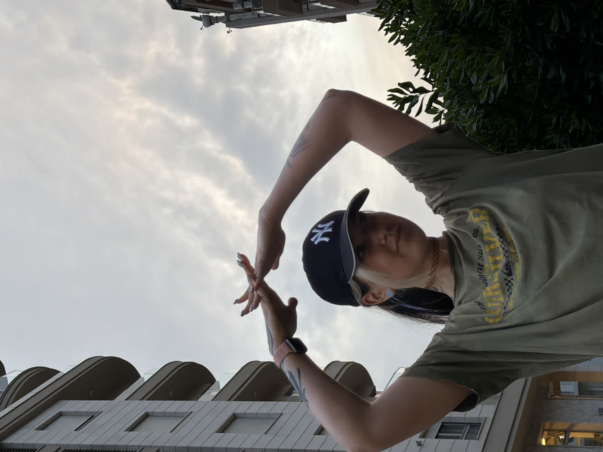 A woman in a green t-shirt and baseball cap stands in Budva, Montenegro, holding her hands to form a heart shape. Her arms are tattooed. She is wearing a smartwatch. In the background are apartment buildings, trees, and a cloudy sky.