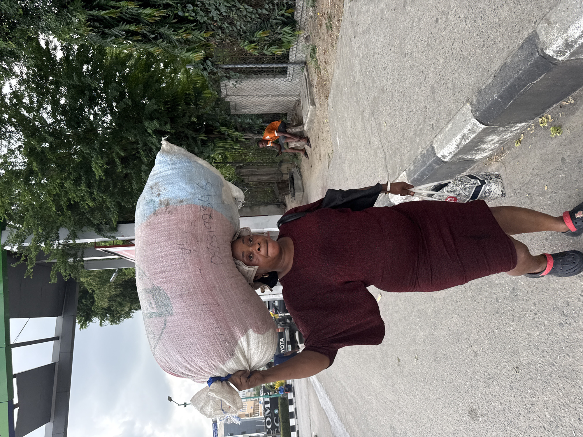 An eye-level, upright shot captures a woman walking along a paved surface, deftly balancing a large, bulky sack on her head. The woman, appearing middle-aged, is dressed in a maroon short-sleeved dress and black Crocs-style sandals with bright red straps. She carries a white plastic bag in her right hand and looks directly towards the camera with a neutral expression.
The sack on her head is substantial, constructed from a woven material, displaying segments of light blue, pinkish-red, and white. Faint black handwritten text, including the numbers "0808219234," is visible on its side. A smaller white bag is tied to the main sack near her left shoulder.
To the left of the frame, a dense canopy of green trees and foliage forms a backdrop. To her right, a paved street or sidewalk with a prominent black and white painted curb is visible. Further back, near a wire fence and bushes, another individual wearing an orange t-shirt and dark shorts is seen squatting or sitting.
In the distance, urban elements such as a grey overhead structure and commercial signage featuring "TOYOTA" and partially obscured "COM" text can be discerned. The sky is bright white, indicating daytime conditions, possibly overcast. The scene depicts a typical street environment in an urban or semi-urban area, consistent with Lekki, Nigeria.