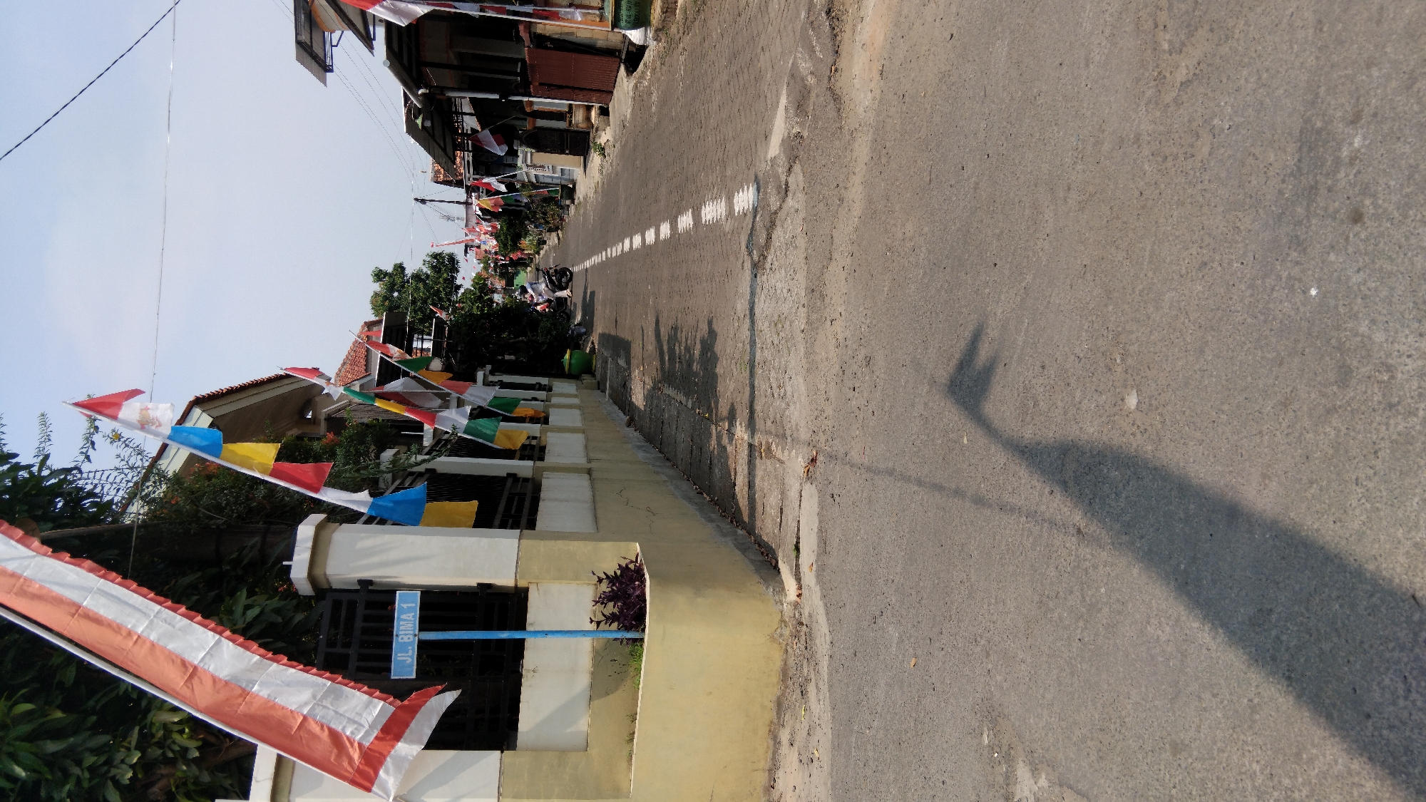 The image shows a street in Pasekaran, Indonesia, decorated with Indonesian flags and banners, suggesting a celebration or holiday. The street is paved with cobblestones and asphalt, with houses lining both sides. There is a blue street sign that reads "JL. BIMA 1". A motorcycle is parked on the side of the road. The sky is clear and bright, indicating a sunny day. The overall scene depicts a residential street adorned for a festive occasion.