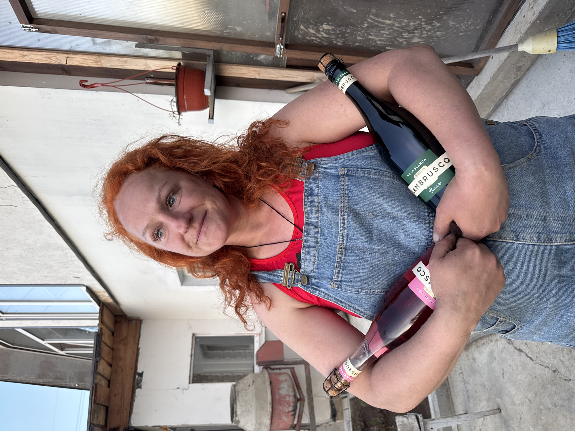 A red-haired, fair-skinned woman wearing blue denim overalls over a red tank top is holding two bottles of Villa Blanca Lambrusco wine. The wine bottles are resting in the crooks of her arms, with a dark green bottle labeled "Bianco" above and a rose-colored bottle below. The background appears to be an outdoor area with building materials, a window, and a hanging clay pot. The location is Chaikovsky, Russia. A cement mixer is visible behind her.