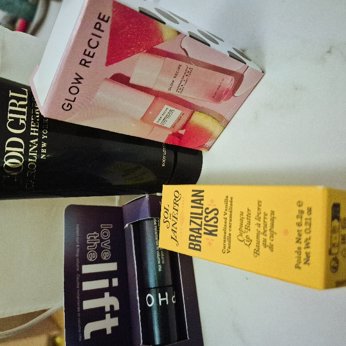 The scene depicts a collection of four beauty and personal care products arranged on a white, smooth surface, likely a countertop or table, in Strasbourg, France. In the center-back is a black tube of "GOOD GIRL CAROLINA HERRERA NEW YORK BODY LOTION" with gold lettering. To its immediate right is a pink and white cardboard box for "GLOW RECIPE" skincare products, featuring a graphic of watermelon slices. To the left of the Carolina Herrera lotion, a dark purple cardboard box with the words "love the lift" inverted is partially open, revealing a dark cylindrical product, likely from Sephora, with "pho" visible on its side. In the foreground, positioned below the Sephora product and the Carolina Herrera lotion, stands a bright yellow rectangular box for "SOL DE JANEIRO BRAZILIAN KISS Caramelized Vanilla Cupuaçu Lip Butter," with "Poids Net 6.2g / Net Wt. 0.21 oz" printed at the bottom. The background is indistinct and light-colored. There are no people present, and no actions or interactions are depicted.