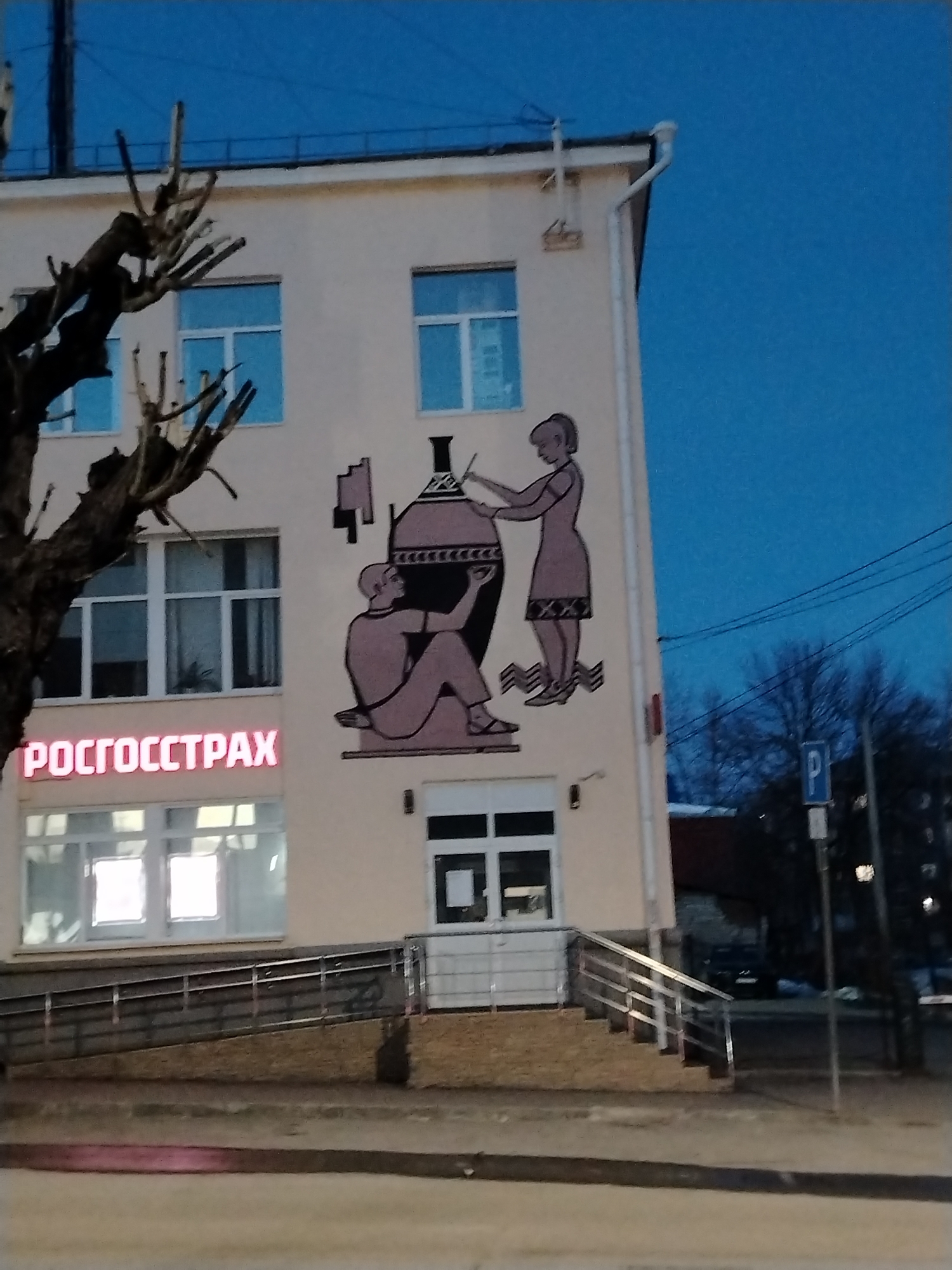 The image shows a three-story building with a mural on its side in Izhevsk, Russia. The mural depicts two figures working on a large vessel or vase in a stylized, possibly ancient Greek or Roman, style. One figure is seated, and the other is standing and appears to be applying or working on the vessel's surface. The building has windows on all three floors, and a sign reading "РОСГОССТРАХ" (ROSGOSSTRAKH), illuminated in red, is visible on the ground floor, indicating the building houses an insurance company. A wheelchair ramp leads to the building's entrance. A bare tree branch is visible on the left side of the frame. A parking sign is visible on the right side of the frame, and the sky is a deep blue, suggesting it is evening or early morning. A set of stairs with metal railings leads to the building's entrance as well.