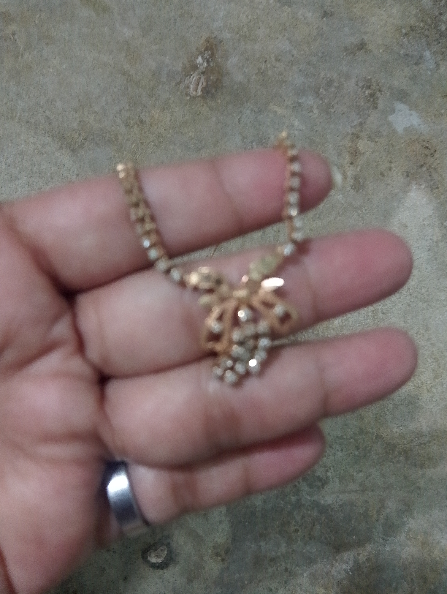 A person's hand, wearing a silver-colored ring, holds a gold necklace with a flower-shaped pendant adorned with small, clear stones. The necklace is positioned over the person's fingers, which are slightly blurred. The background appears to be a rough, textured surface, possibly concrete or stone, with some small debris. The image is taken in a dimly lit indoor or outdoor setting. The city of Serbelawan, Indonesia, is not discernible from the visual information provided in the image.