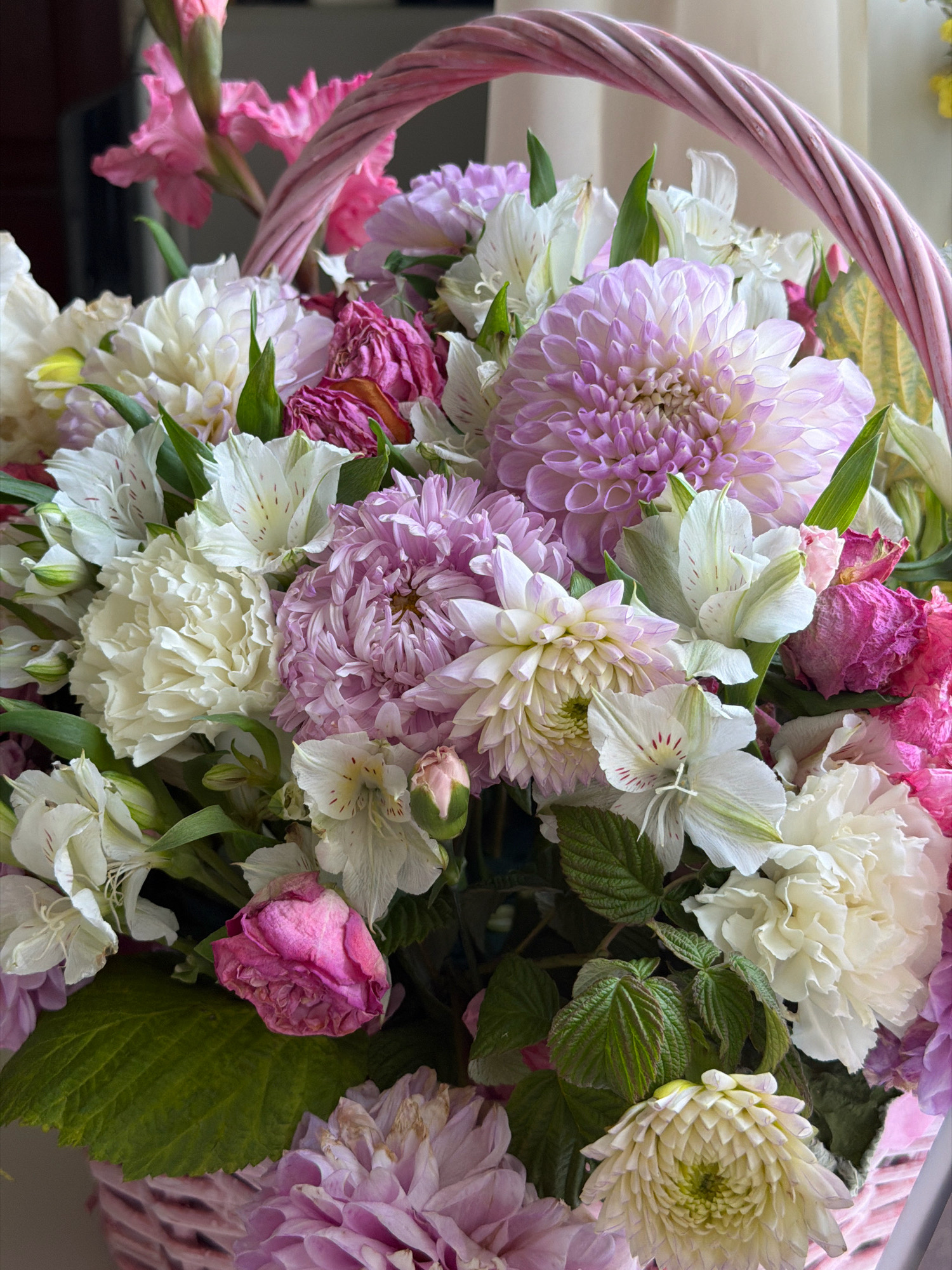 A close-up, eye-level shot captures a wicker basket overflowing with a vibrant arrangement of flowers. The basket itself has a pink, twisted rope handle. The flowers are a mix of pink, white, and light purple hues, with varying textures and petal formations. Prominent are large, fluffy white and pink dahlias, interspersed with smaller white alstroemeria and delicate pink gladiolus blooms. Some of the pink flowers appear to be wilting, showing dried petals. Green foliage peeks through the colorful blossoms. The background is blurred, suggesting an indoor setting with diffused light. The image does not contain any people. The location in Бабініцкі сельскі Савет, Belarus, is not discernible from the visual information provided.