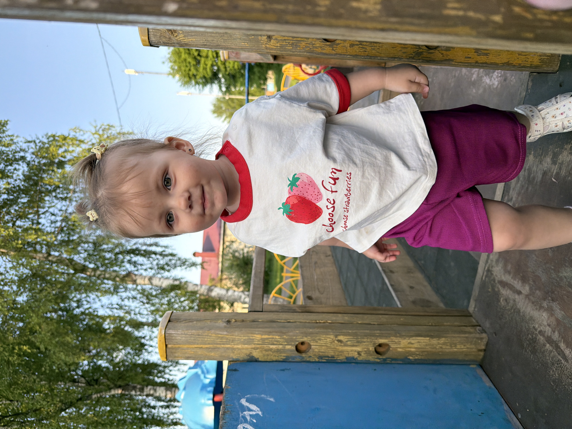 A child stands on a playground structure. The child is fair-skinned with light brown hair tied in pigtails. She wears a white t-shirt with red trim and strawberry designs, and purple shorts. The structure is made of wood and metal and painted blue and yellow. Trees and a grassy area are visible in the background. The city of Reshetnikovo, Russia, is not discernible from the provided image.