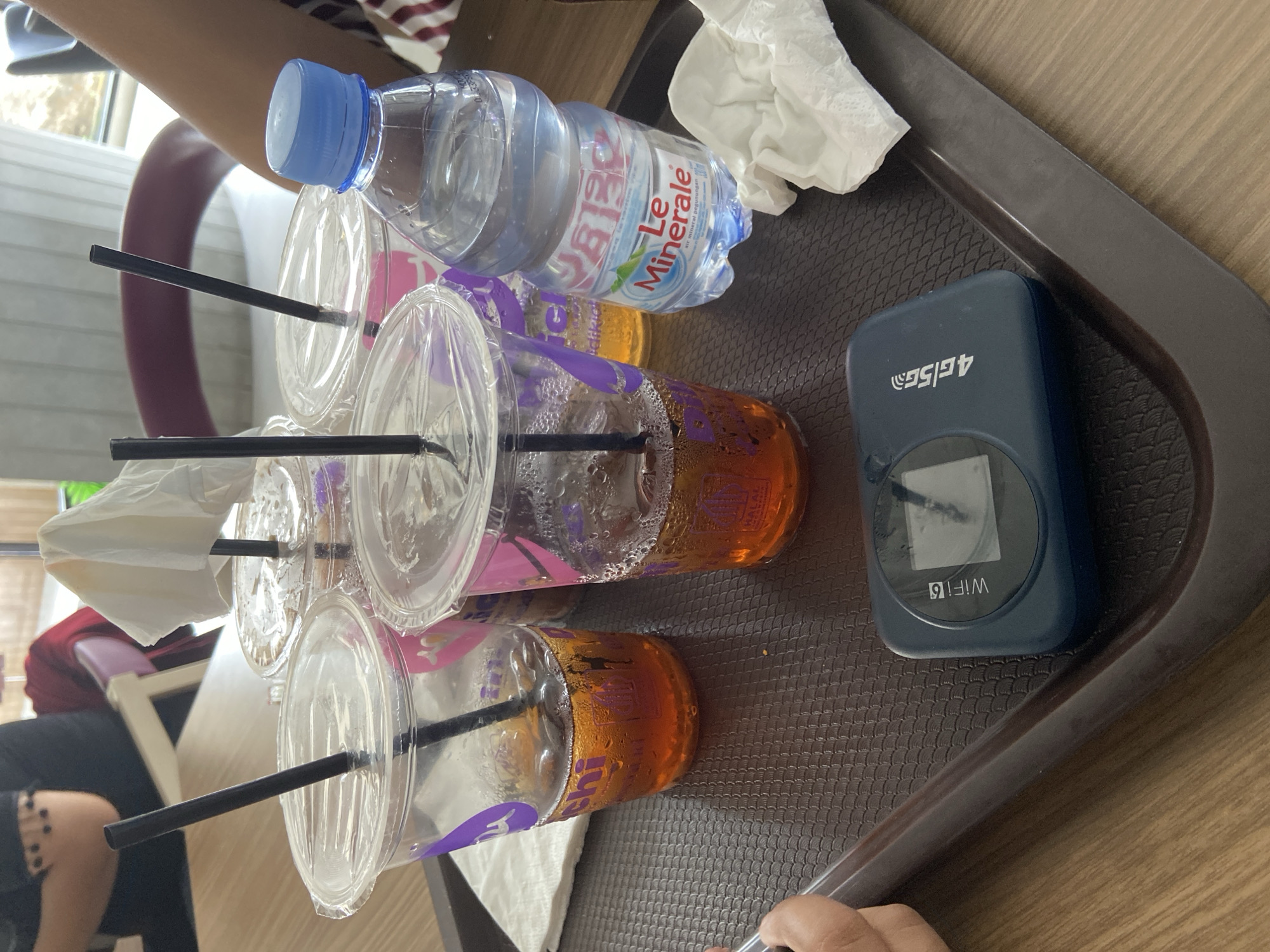 A dark brown tray holds four plastic cups of iced tea, a bottle of "Le Minerale" mineral water, two crumpled white napkins, and a dark blue portable Wi-Fi hotspot device. The cups are partially filled with amber-colored iced tea and have clear plastic lids with black straws. The bottle of water has a blue cap and a label with green and red text. The portable Wi-Fi device displays the text "WiFi" and "4G|5G". The scene appears to be indoors, possibly at a cafe or restaurant in Bago, Indonesia, with a wooden table surface visible. In the background, parts of individuals are visible, including the arms and shoulders of someone wearing a black top with decorative elements on the shoulder and another person in a red garment.