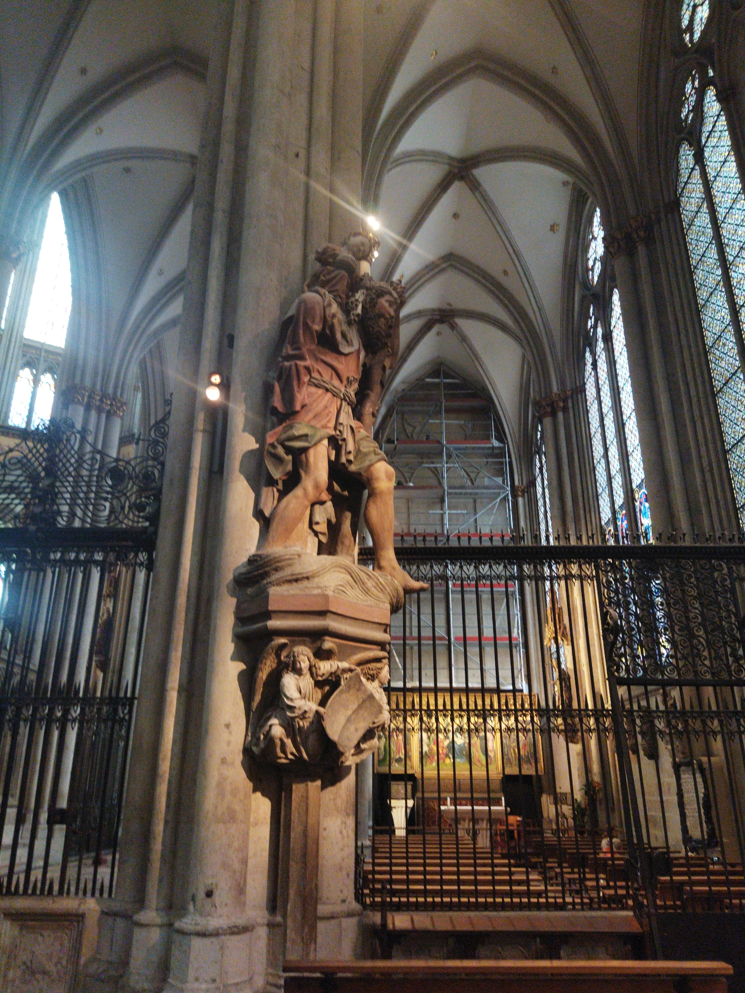 This image shows an interior view of a Gothic cathedral, likely Monheim am Rhein's parish church of St. Gereon, given its architectural style and the presence of a prominent sculpture. A large, stone sculpture of two figures, one carrying the other, stands on a pedestal in the foreground, dominating the left side of the frame. The sculpture is detailed, with the figures appearing to be in motion. Behind the sculpture, an ornate iron fence separates the viewer from the rest of the nave. Sunlight streams through tall, arched windows on the right, illuminating the space and casting long shadows. In the background, scaffolding is visible, suggesting ongoing renovation or construction work. Rows of pews are arranged in the nave, and a large altarpiece is partially visible. The architecture features ribbed vaults, pointed arches, and stone columns, characteristic of Gothic cathedrals. No people are clearly identifiable in the image.