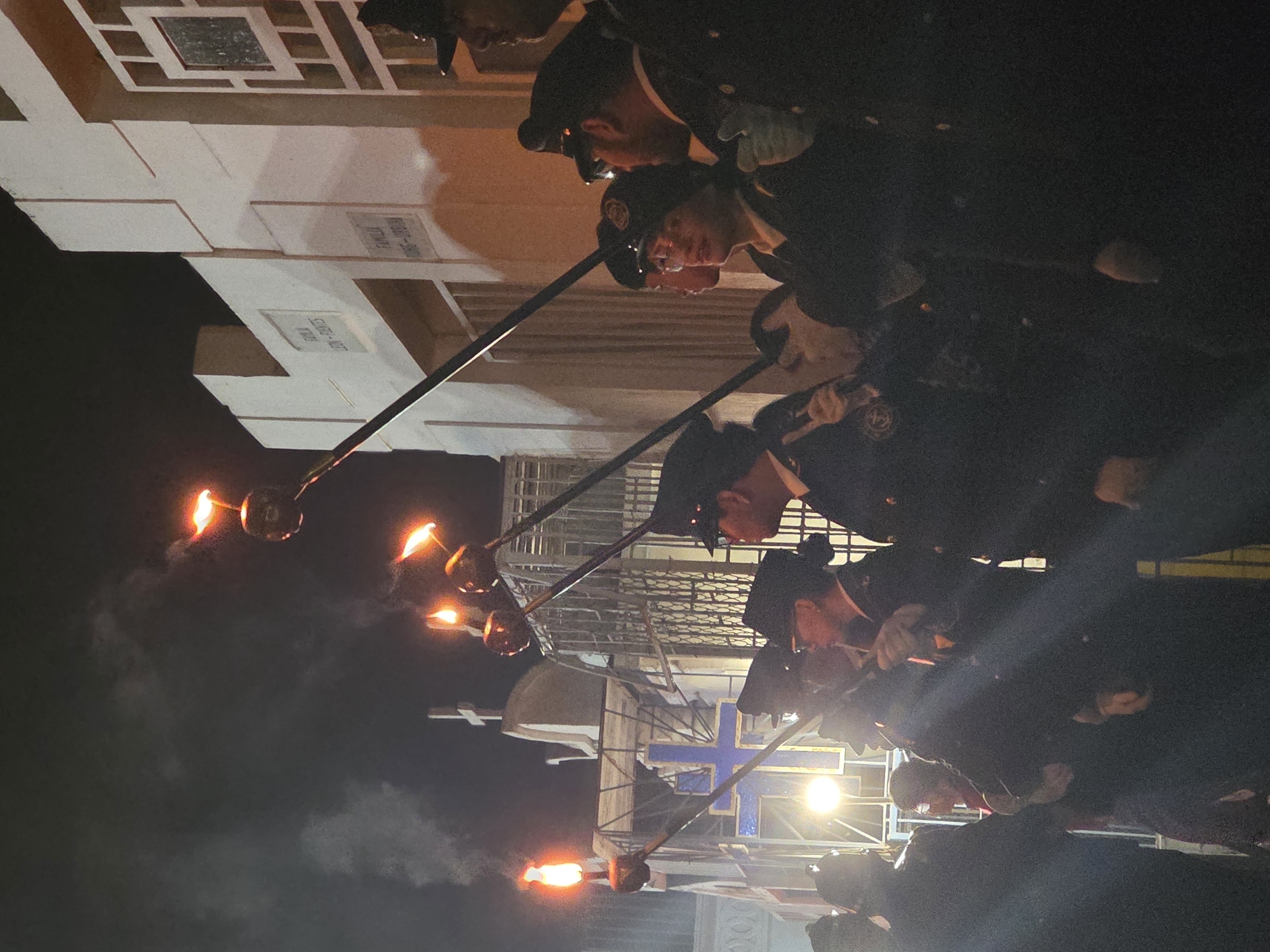 The image shows a procession at night in Iquique, Chile. The camera angle is low, looking up at a group of men in dark uniforms and caps, each holding a long pole with a flaming torch at the end. They are walking past crypts in a cemetery. A prominent blue cross is visible in the background. The atmosphere appears solemn.