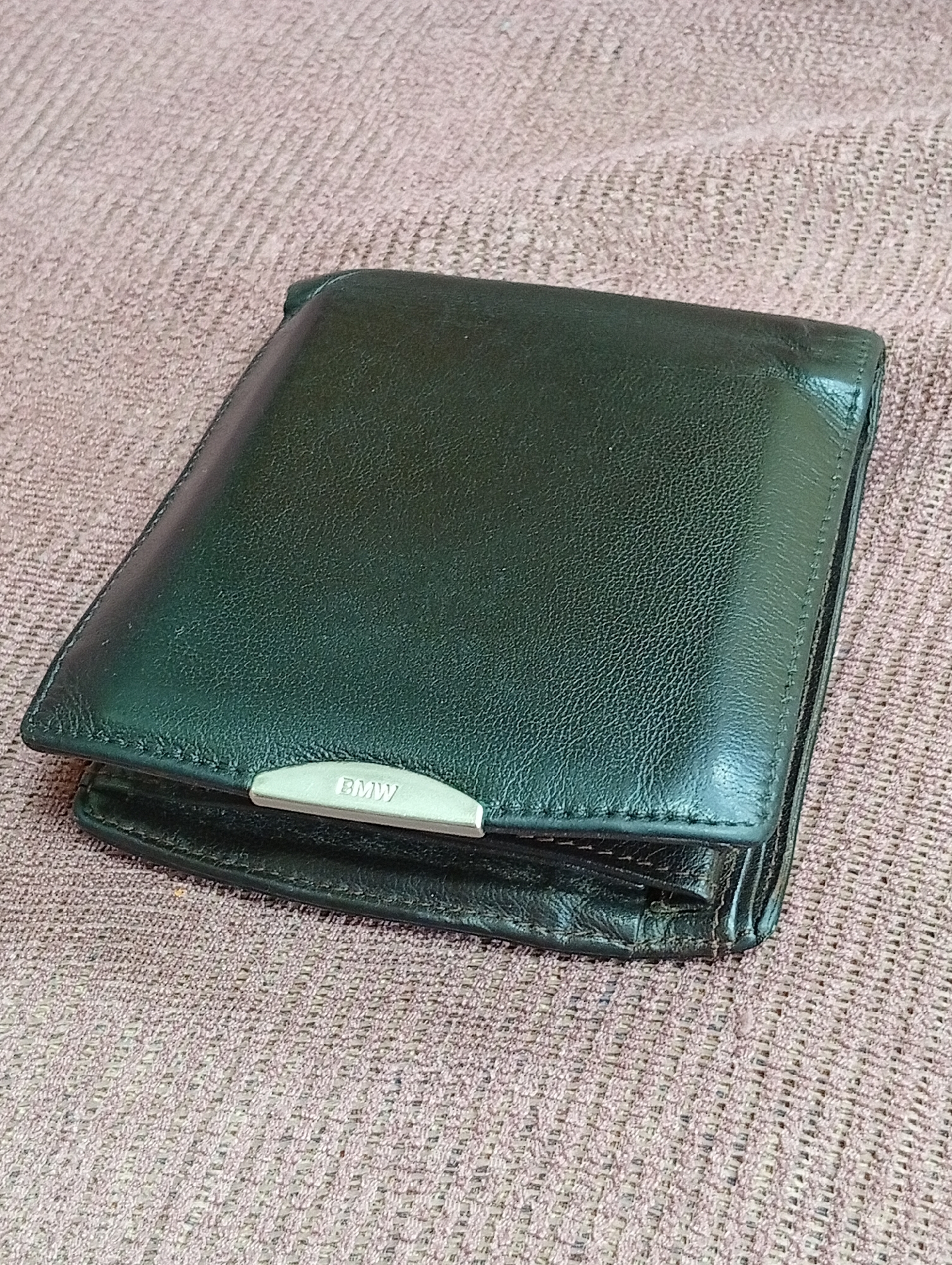 A dark green leather bi-fold wallet with a silver-toned metal accent featuring the "BMW" logo is resting on a textured, pinkish-brown fabric surface. The wallet is closed and appears slightly worn, with visible creasing and some scuffing on the edges. The fabric surface has a woven texture, suggesting it could be a tablecloth, upholstery, or a similar textile. The lighting creates some shadows and highlights on the wallet, emphasizing its material and form. The image is a close-up, focusing on the wallet and its immediate surroundings. The location in Крыўлянскі сельскі Савет, Belarus is not discernible from the image.