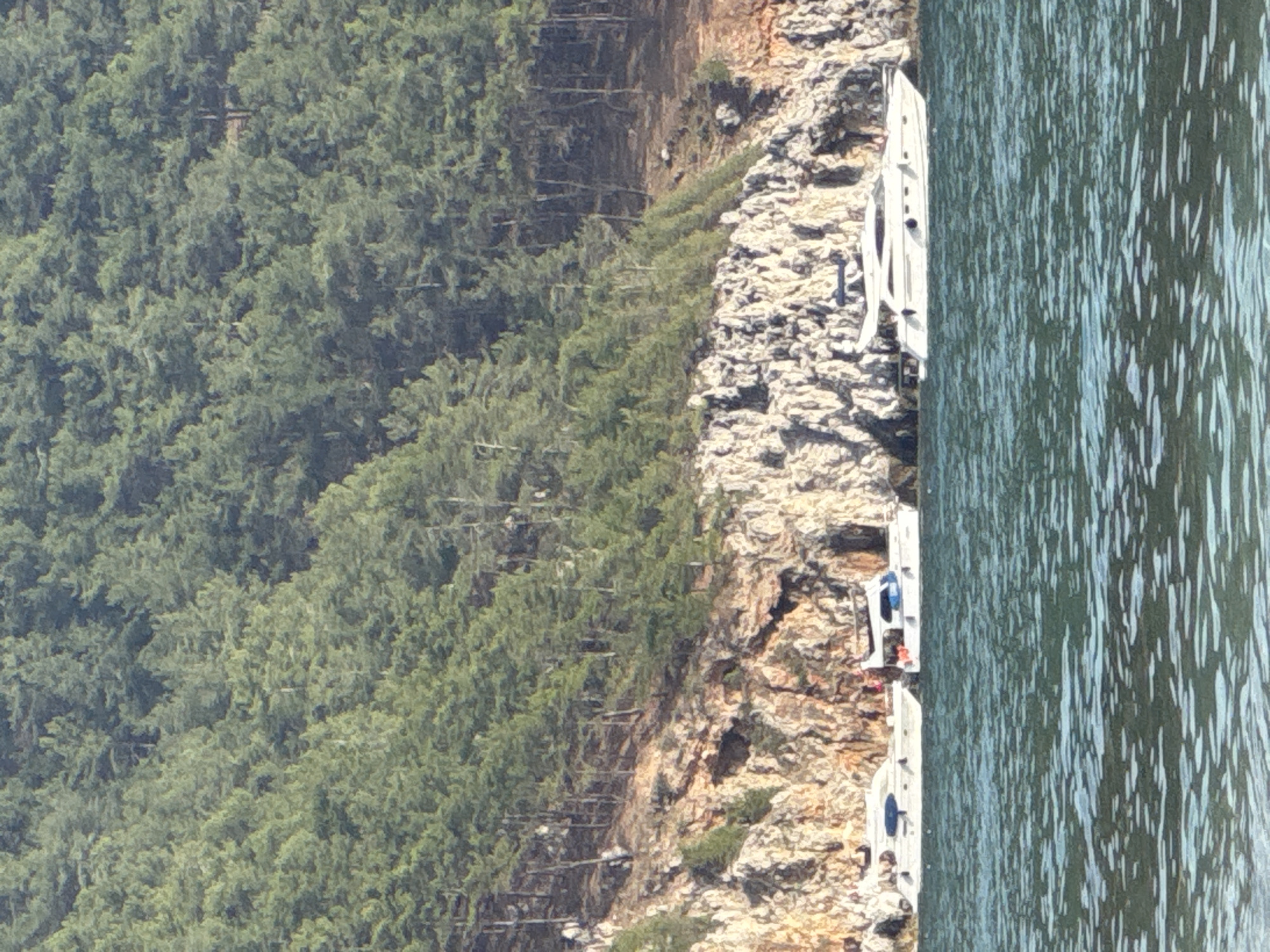 The image depicts a scene in Alanya, Turkey, showing a shoreline with lush green vegetation meeting rocky cliffs. Several small white boats are docked along the rocky edge of the cliffside, with the sea directly adjacent. People appear to be near the boats. The water shows slight rippling, indicating a mild breeze.