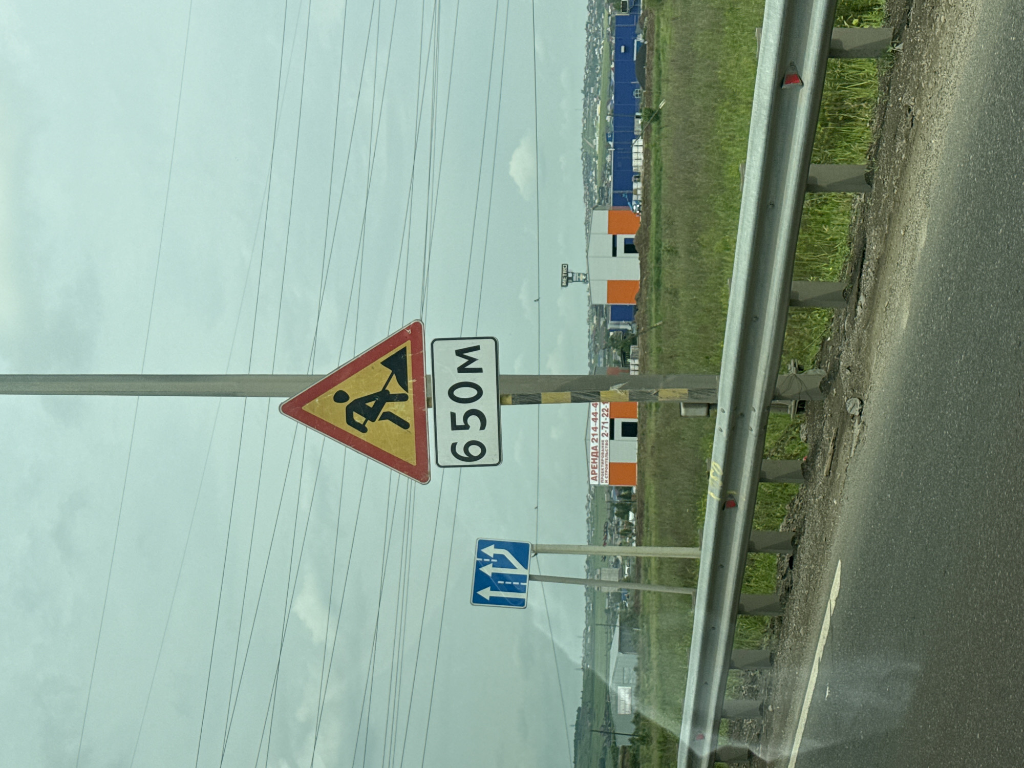 The image shows a roadside scene with several traffic signs in Solontsy, Russia. In the foreground, a road with a metal guardrail runs along the right side. Beyond the guardrail is a grassy field. In the background, there are buildings, including some under construction with orange and white panels, and a city skyline. The sky is overcast.
The traffic signs are mounted on poles. The topmost sign is a yellow triangle with a red border, depicting a person with a shovel, indicating road work ahead. Next to it is a rectangular sign with the number "650" in black on a white background, likely indicating the distance to the road work. Below these, a blue rectangular sign shows two lanes merging into one, with arrows indicating traffic should move to the right.
A smaller sign with text is visible on a pole in the background, appearing to be an advertisement. It reads "АРЕНДА 214-44-44" and "И СТРОИТЕЛЬСТВО 271-22-1". This suggests the rental and construction business.
The scene appears to be captured during the daytime, with the cloudy weather creating a diffused light. The overall impression is a functional roadside environment with ongoing construction or maintenance work.