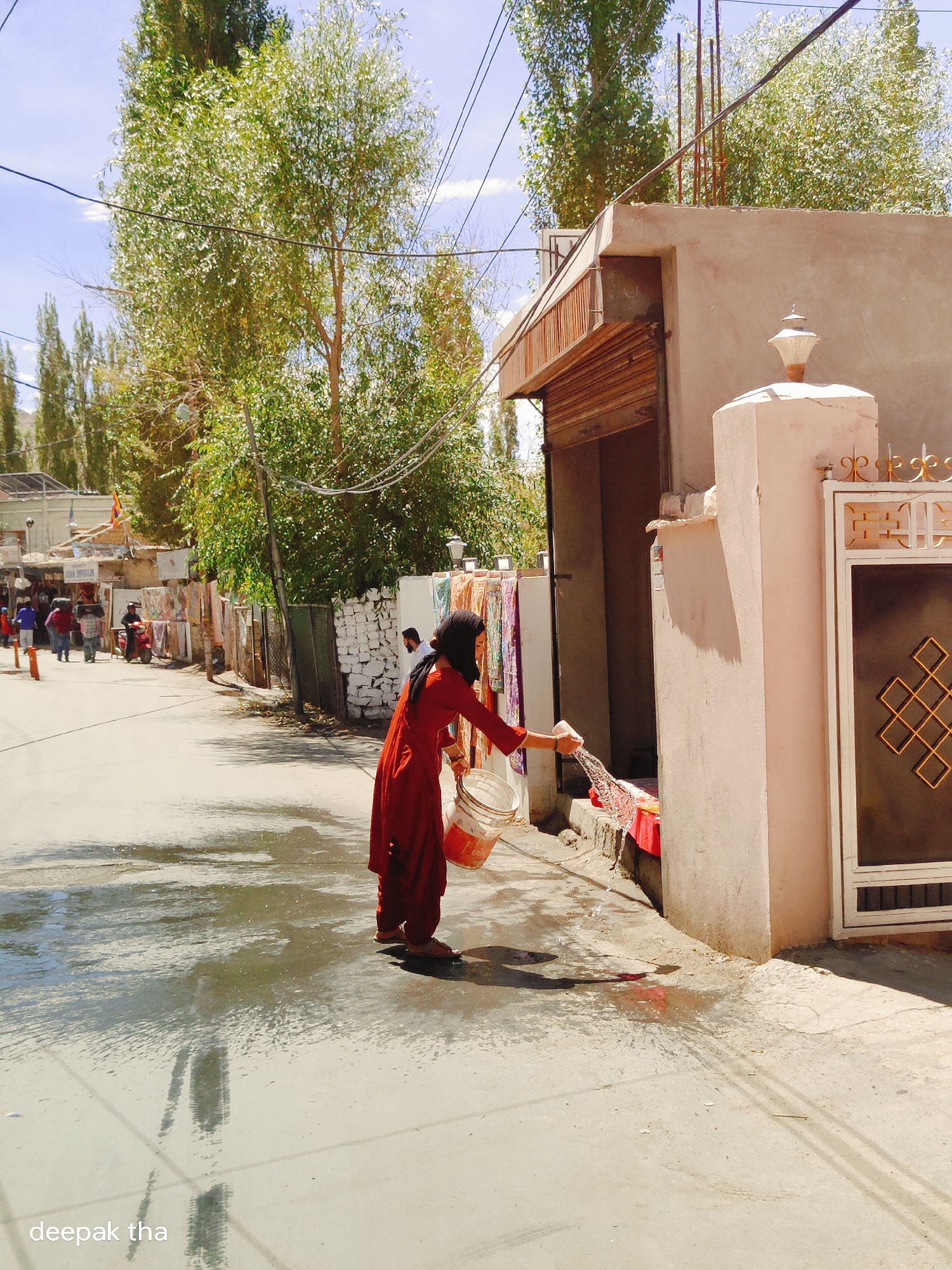 In Leh, India, a woman wearing a red shalwar kameez and a black headscarf is captured in a candid moment, outdoors. She is standing on a wet, paved surface, likely a street or pathway, in front of a building. In her right hand, she holds a white plastic bucket, from which she is pouring water with a string or cloth. The water is creating a puddle on the ground. Behind her, to the left, a line of shops with various goods displayed can be seen. The buildings are typical of the region, with flat roofs and simple architecture. Tall trees with green foliage line the background, and the sky is a clear blue with some wispy clouds. The overall scene depicts a common, everyday activity in the environment of Leh, a city located in the Indian Himalayas.