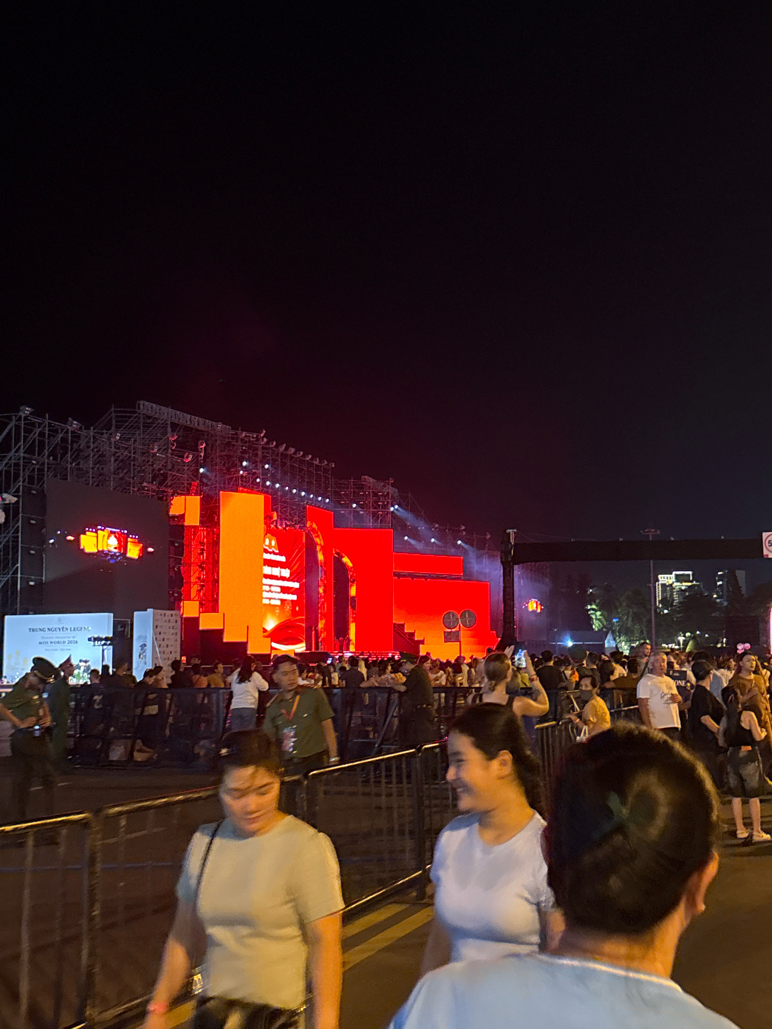 This image depicts an outdoor evening event in Nha Trang, Vietnam, with a large stage set up in the background. The stage is illuminated with bright red and orange lights, and features a large screen displaying content, as well as structural elements. A crowd of people is gathered in front of the stage, separated from the foreground by a metal barricade.
In the foreground, three individuals are visible. A woman in a light-colored t-shirt and dark pants is walking towards the left of the frame, her body angled away from the camera, and she is looking to her left. To her right, another woman in a light blue t-shirt is looking towards the right of the frame with a slight smile. Her face is turned towards the camera. In the immediate foreground, the back of a person's head and shoulders is visible, facing away from the camera and towards the event. They are wearing a light blue shirt.
Further back, near the stage, several individuals are standing and walking. Some appear to be security personnel in uniforms. Signs are visible near the stage, one of which reads "TRUNG NGUYEN LEGEND" and "MISS WORLD 2026". The environment appears to be a public space, possibly a park or a plaza, given the presence of trees and distant buildings visible on the right side of the frame. The overall atmosphere suggests a concert or a large public gathering.