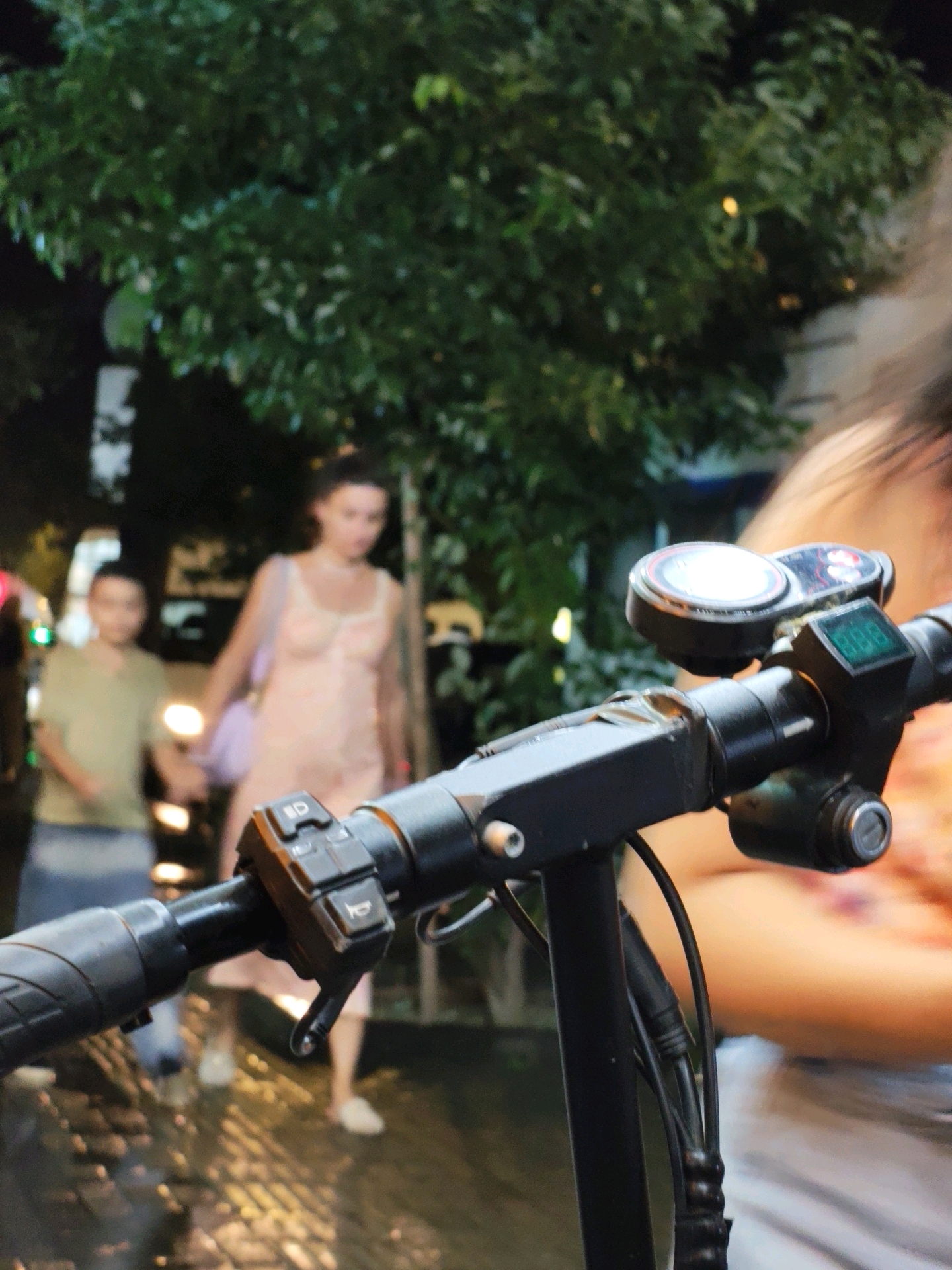 A low-angle, dynamic, first-person perspective shot captures the handlebar of a black electric scooter as it moves along a wet, cobblestone street at night. The scooter's control panel, featuring a digital display and buttons, is in the foreground, with visible wiring. In the background, a young woman in a light-colored dress and a young man in a light green shirt are walking away from the viewer. The woman is carrying a purple bag. Lush green trees and the blurry lights of the city line the street, suggesting an urban environment. The wet cobblestones reflect the surrounding lights, indicating recent rain. The scene appears to be set in Batumi, Georgia, known for its coastal location and vibrant nightlife, especially around the area of the Batumi Boulevard.