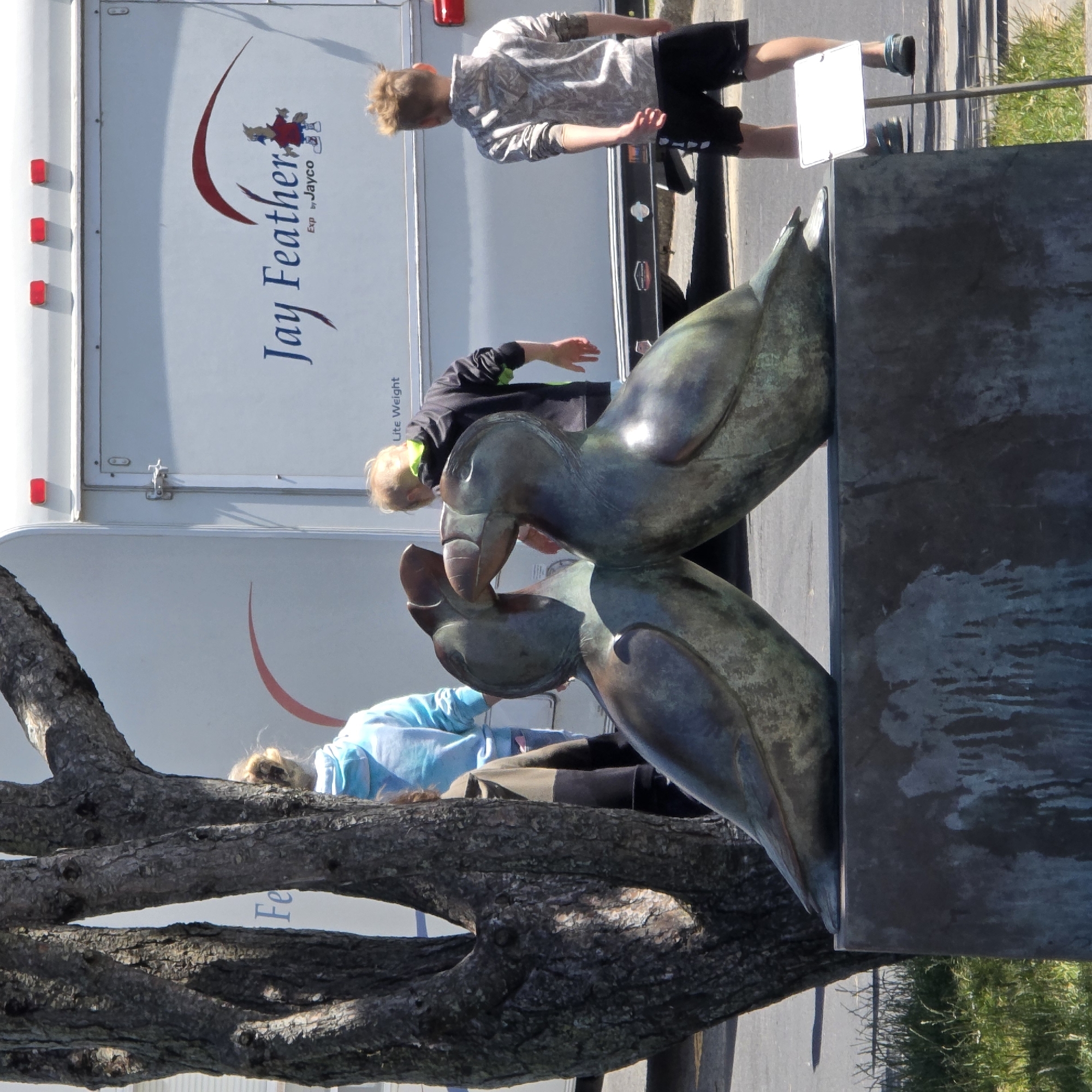 A bronze sculpture of two puffins, with their heads facing each other, is located on a stone pedestal in front of a white trailer with the text "Jay Feather" and a cartoon bird logo. In the background, a tree trunk is visible on the left. Three people are present: a woman wearing a light blue hooded sweatshirt, a boy in a black and green hooded sweatshirt, and another boy in a camouflage hooded sweatshirt and black shorts. The scene is outdoors on a sunny day, with grass and asphalt visible in the foreground and background, respectively. The image appears to have been taken in Cannon Beach, Oregon, United States of America, known for its distinctive rock formations and puffin populations.