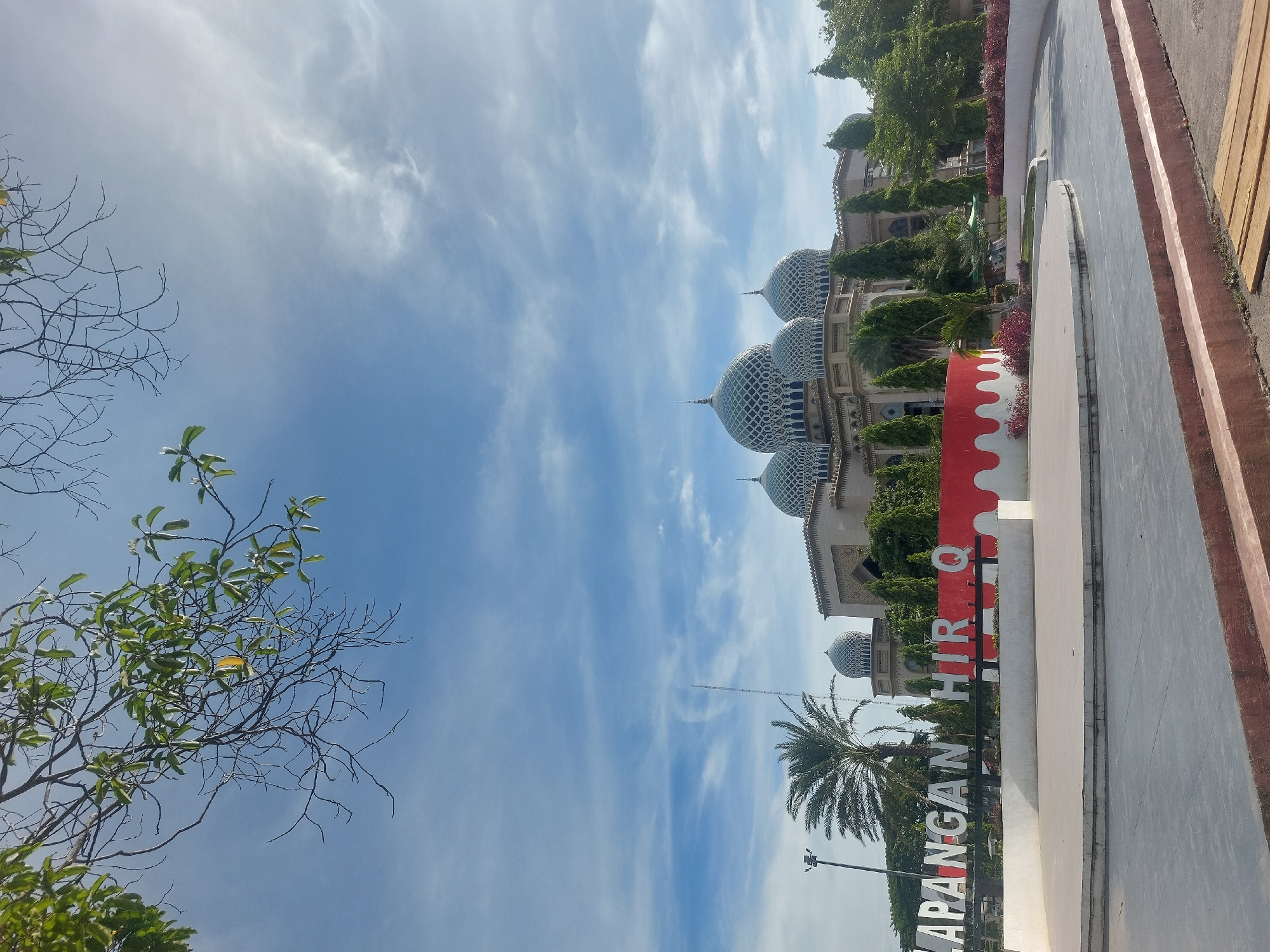 The image captures the Lapangan Hiri Q, a public square in the city of Lhokseumawe, Indonesia, on a bright, partly cloudy day. Dominating the background is a grand mosque with multiple domes adorned with intricate patterns, characteristic of Islamic architecture. In the foreground, large white letters spelling out "LAPANGAN HIRI Q" stand prominently, partially obscured by a red and white decorative border. A palm tree and other lush greenery surround the plaza, contributing to the tropical ambiance. The plaza itself features a paved area with raised platforms and seating, suggesting it is a space for gatherings and events. The sky is a vibrant blue, with scattered white clouds, and a few branches of a tree with green leaves are visible in the upper left corner, framing the scene.