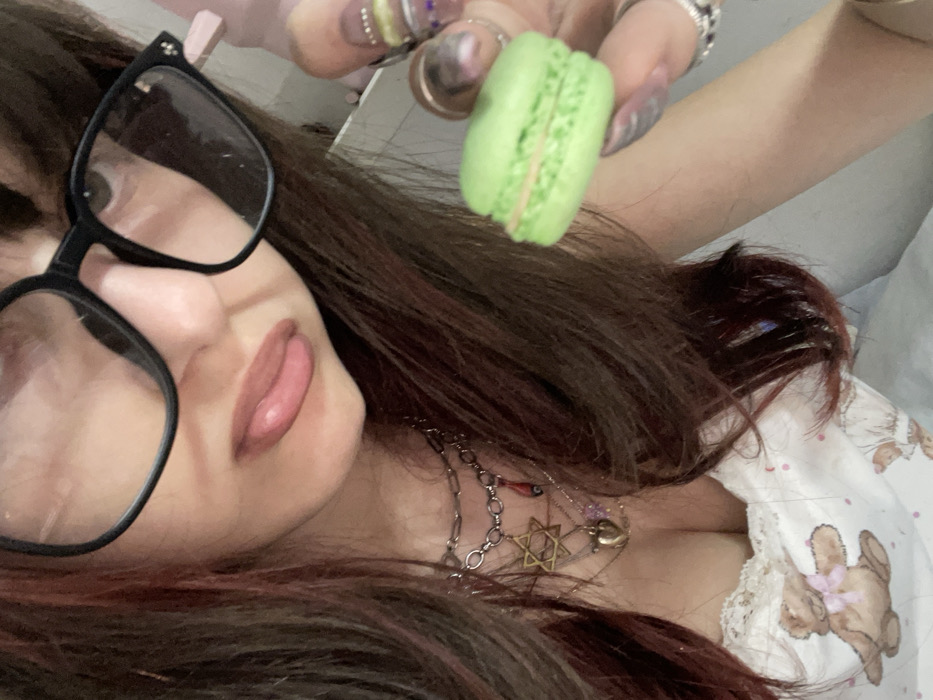 A person wearing black glasses and multiple necklaces, including one with a Star of David pendant, holds a green macaron. The individual has long, brown hair with red highlights and is wearing a white top adorned with teddy bear patterns and lace trim. The person's fingernails are decorated with a variety of embellishments and rings. The perspective is close-up and slightly angled.