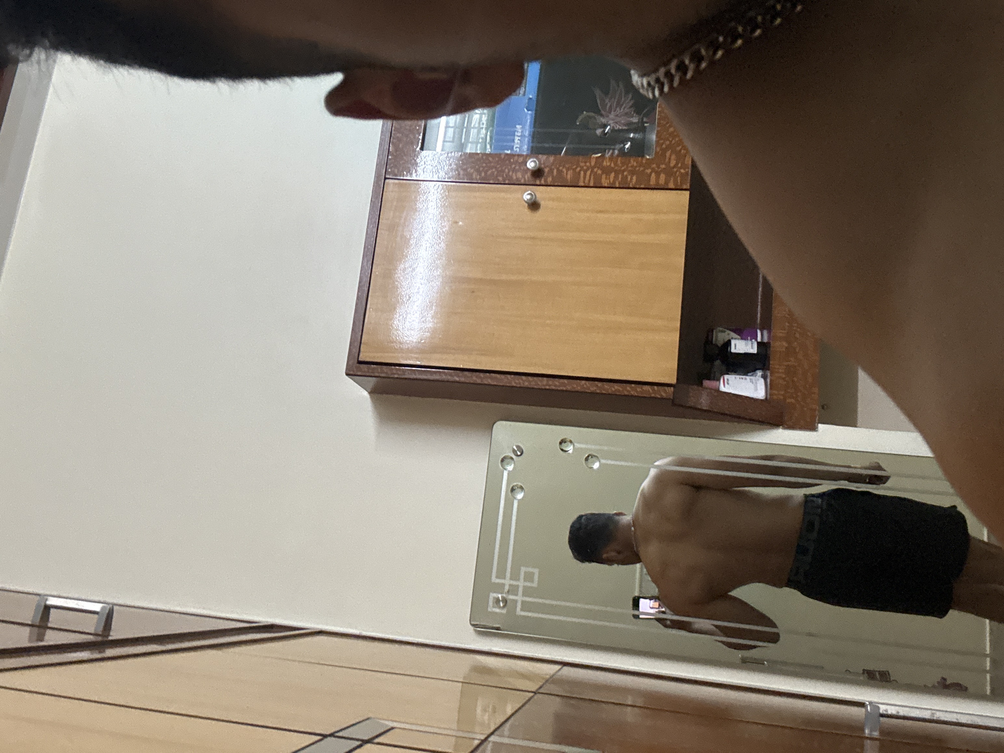 The image presents an indoor scene, likely a personal room such as a bedroom or dressing area, captured from a first-person perspective. In the upper left foreground, the top of a person's head with dark hair is visible, along with their arm adorned with a silver-colored chain or bracelet, indicating they are holding the camera themselves.
The central subject, a male with a muscular physique, is prominently featured in the reflection of a decorative wall mirror. He is shirtless, wearing dark shorts or underwear, and is actively taking a selfie using a smartphone held in both hands. The reflection shows his back and side profile, highlighting his developed shoulders and back muscles. The smartphone screen, displaying the camera interface, is visible in his hands.
The mirror itself is rectangular, featuring beveled edges and decorative etched patterns along with what appear to be clear glass or crystal studs. It is mounted on a light-colored wall. Above the mirror and to the right, a wooden wall-mounted cabinet or shelf unit is visible. This unit has a lower section with a wooden door and an upper section with a glass front, behind which some indistinct items are stored. The word "SYSTEM" can be faintly discerned on one of the items inside the glass cabinet. The floor at the bottom of the frame is tiled, with a light, reflective surface. The overall lighting suggests an indoor setting with artificial illumination. The scene captures a moment of self-documentation, likely focusing on physical appearance.