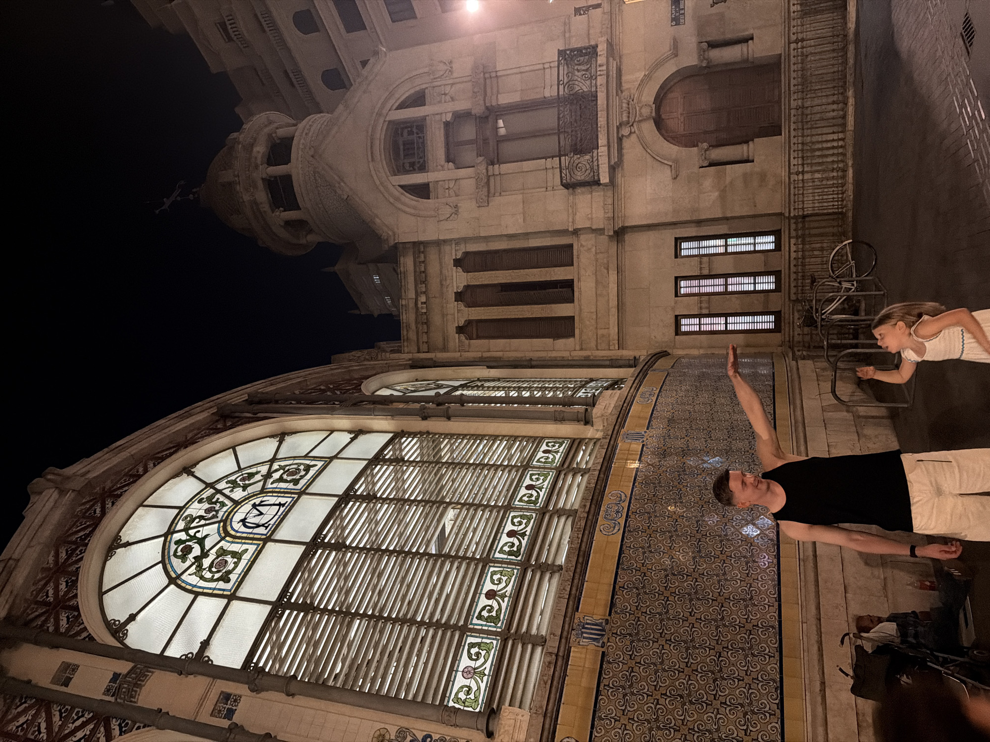 The image captures a nighttime scene in front of the Mercado de Colón, a modernist architectural building located in Valencia, Spain. On the left, a man wearing a black tank top and white shorts stands facing right, with his right arm extended, appearing to point or gesture. Beside him and slightly to his right, a young girl in a white dress looks up towards the man's hand. Behind them, the ornate facade of the market is visible, featuring large arched windows with decorative stained glass and intricate tilework. To the right of the market building, another stone structure with arched windows and a balcony is illuminated. A bicycle is parked in the foreground, and a stroller is partially visible to the far left. The overall lighting suggests it is after dark, with artificial lights illuminating the buildings and the immediate area.