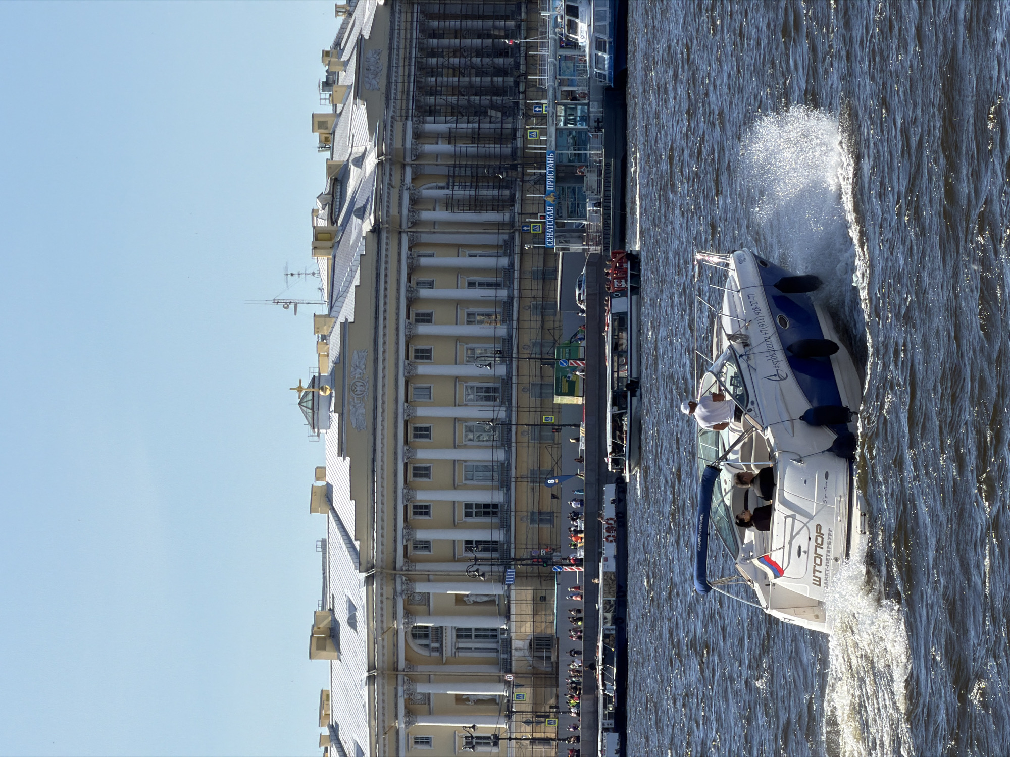 A pleasure boat, with "ШТОПОР" and "SAINT-PETERSBURG" written on its stern, speeds across dark, choppy water, creating a large white spray. The boat has a blue canopy and is occupied by at least three people, two seated and one standing near the helm. In the background, a long, pale yellow neoclassical building with numerous columns and windows is visible. Scaffolding covers portions of the building, suggesting ongoing renovation. A clear blue sky stretches above. Along the waterfront in front of the building, a crowd of people are gathered, and several other boats, including larger tour boats and a smaller speed boat, are visible on the water. Signs with Russian text, such as "СЕНАТСКАЯ ПРИСТАНЬ" (Senate Embankment Pier), are present. The scene is likely Saint Petersburg, Russia, given the architectural style and the presence of the Russian flag on the boat and the text "SAINT-PETERSBURG."