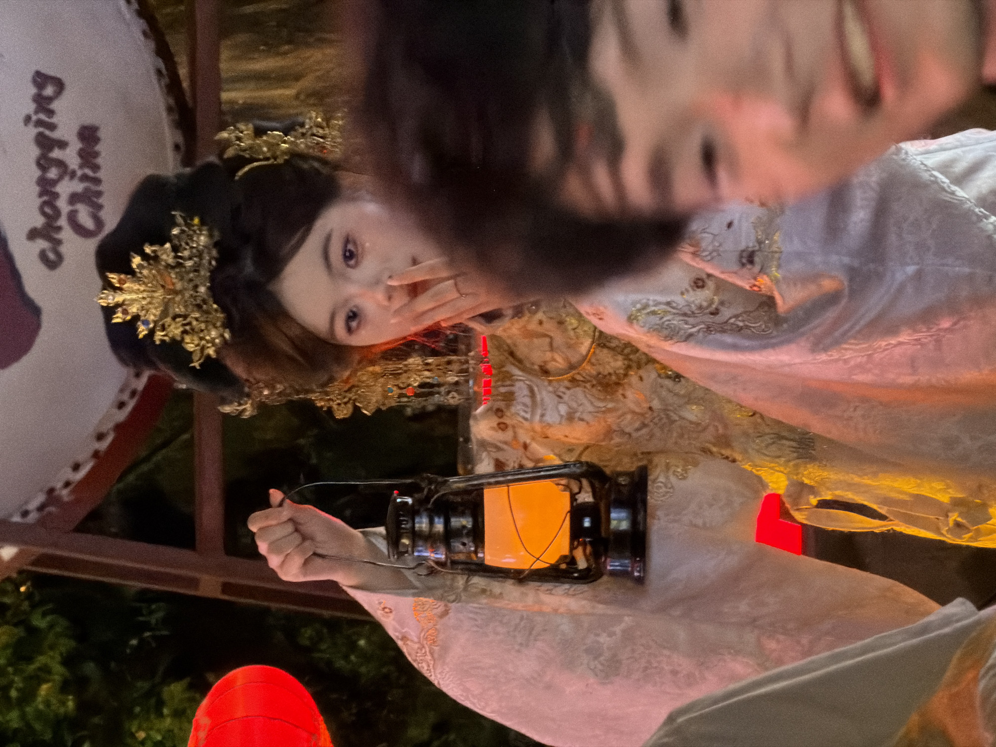 A young woman, dressed in traditional Chinese attire with an elaborate golden headdress, stands in what appears to be a festive or historical reenactment setting. She holds a black, vintage-style lantern with a glowing orange light in her left hand. The lantern's light illuminates her pale face and the intricate details of her white and gold embroidered robe. Her right hand is raised to her mouth, with a delicate ring on one finger. In the foreground, a blurred figure, possibly another attendee, is visible, their face out of focus. Behind the woman, a large drum with "chongqing China" written on it is partially visible, suggesting the location might be Chongqing. The background is dark, with hints of red lanterns and greenery, creating a warm, somewhat mysterious atmosphere. The image captures a moment of cultural performance or celebration in Yuzhong District, China.