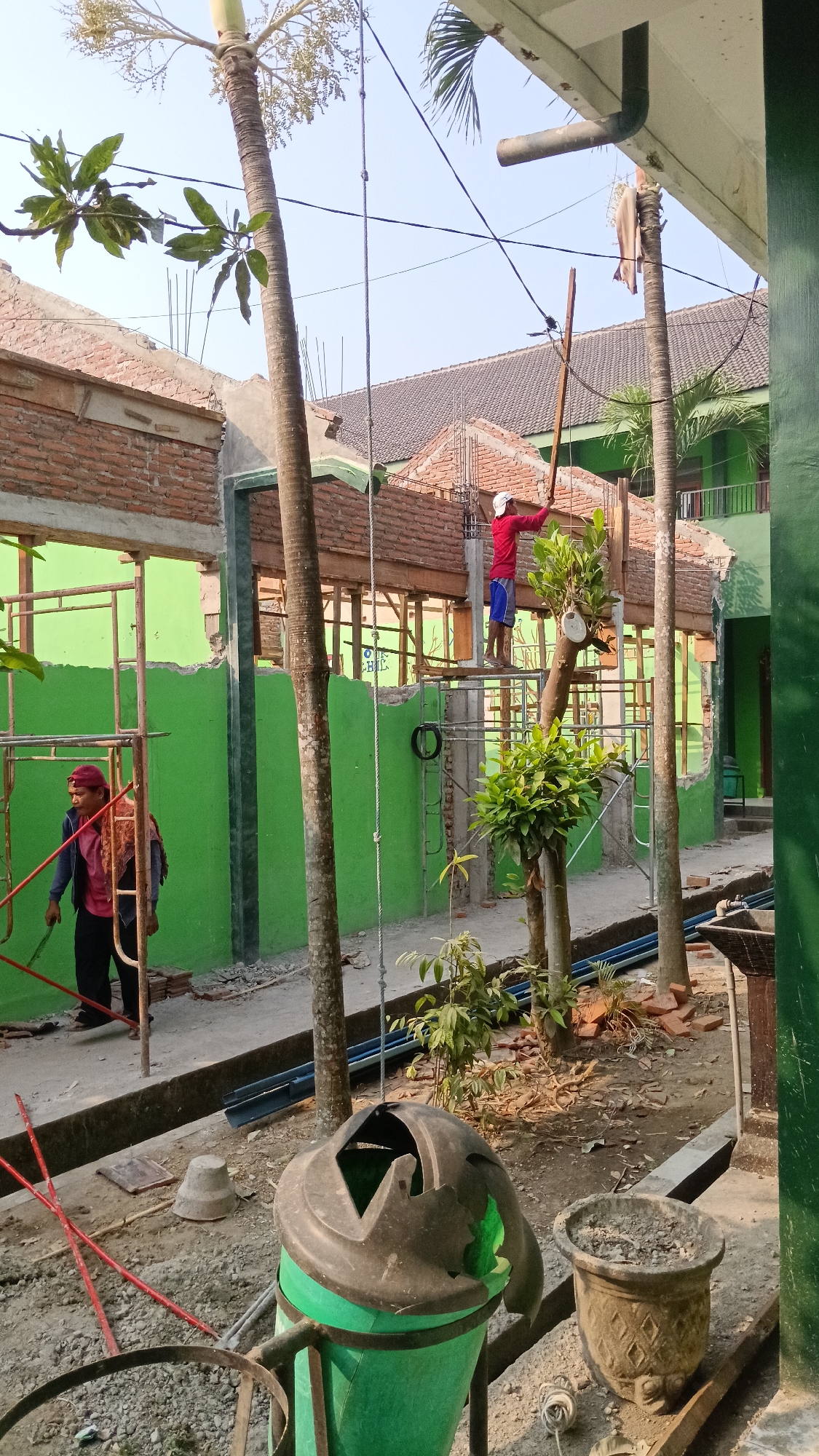 The image depicts an outdoor construction or renovation scene in Branggahan, Indonesia. Two individuals are present. One person, wearing a red shirt and blue shorts, stands on scaffolding, holding a wooden pole and appearing to work. The other person, wearing a maroon cap, a maroon shirt, and dark pants, stands on the ground to the left, holding what appears to be a tool or a piece of rebar.
The environment is characterized by a building undergoing repair, with exposed brickwork and scaffolding. The walls of the building are painted bright green, but sections appear to be damaged or under construction. Several palm trees and other foliage are scattered throughout the scene, with one particularly large tree trunk dominating the central foreground. In the foreground, there is a green trash can with a damaged black lid, and a decorative planter. The ground is covered with gravel, dirt, and scattered construction debris, including red poles. The sky is clear and bright, indicating daytime.