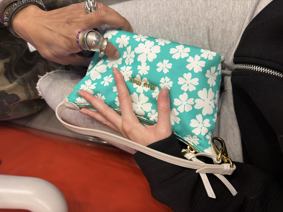 The image displays a close-up view of two individuals' upper legs and hands, centered on a Miu Miu clutch bag, within an indoor environment in Paris, France. A fair-skinned hand, with neat, short nails, is visible on the left, resting on a small, rectangular clutch bag. This bag is turquoise with a repeated white floral pattern and features a gold-toned "MIUMIU" logo on its front, a silver zipper along its top edge, and a light beige wrist strap secured by a gold-colored lobster clasp. The individual's arm is clad in a black long-sleeved garment, and their lap is covered by grey sweatpants. To the right, a darker-skinned hand is positioned near the bag. This hand is heavily accessorized, featuring a large, multi-segmented silver-toned ring on the thumb, a ring with multiple small purple stones on the index finger, a dark ring on the ring finger, and an intricate silver-toned ring on the pinky. A dark, possibly studded, bracelet is visible on the wrist. The individual's lap is also covered by grey sweatpants, and a camouflage-patterned fabric is visible on the upper thigh. The background on the left consists of an out-of-focus bright orange-red surface against which a white, curved object is partially seen. The two hands are in close proximity, with the fair-skinned hand lightly touching the bag, and the darker-skinned hand resting nearby, implying a shared space or interaction.