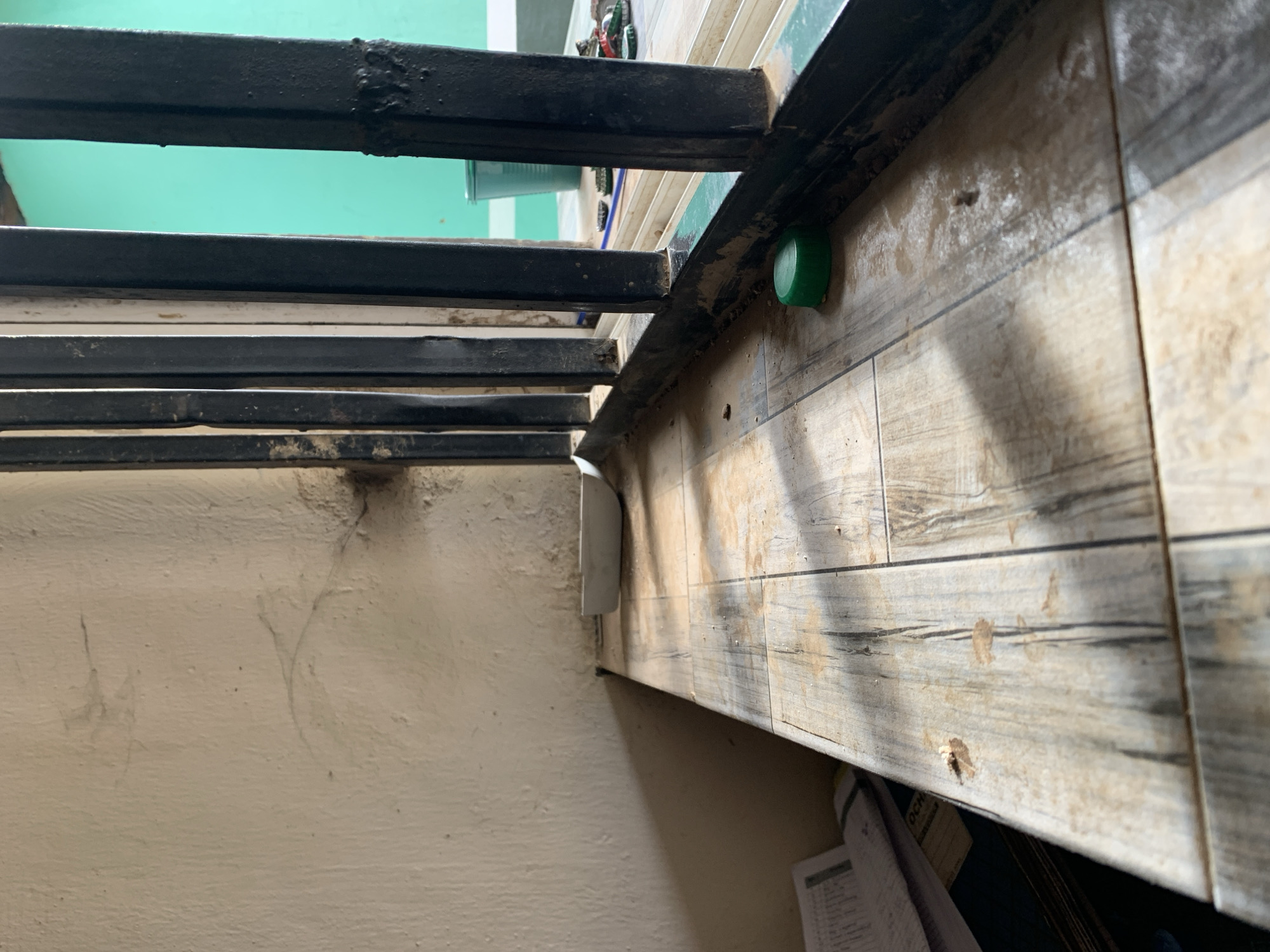 This forensic description details an indoor scene, likely a room, facing outwards through a barred opening, located in the city of New Stadium, Nigeria.
The dominant feature is a tiled ledge or sill extending horizontally from the left, composed of light brown and grey tiles arranged in a wood-grain pattern. This surface is visibly dusty and soiled with dirt particles and several distinct brown stains, suggesting dried residue. In the upper left quadrant of the ledge, a small, white, shallow scoop or dish rests. In the lower right foreground, a bright green plastic bottle cap lies on the tiled surface.
To the right of the ledge, a series of vertically oriented, black metal security bars are installed. These bars are thick and show signs of wear, including rough paint texture and areas of apparent corrosion or heavy weld marks, particularly on the outermost right bar. The bars cast distinct shadows onto the tiled ledge, indicating a light source from outside.
Beyond the bars, the exterior environment is visible. A prominent wall painted a vivid turquoise or teal green forms the background. To the left of this green wall, a lighter-colored vertical structure is partially visible. On an exterior surface, roughly at the level of the tiled ledge, a green plastic bucket or container is present, partially obscured by the bars. Scattered to the right of the bucket are several discarded bottle caps or small lids, some red and some metallic, lying on the ground or an external sill.
To the left of the tiled ledge, a textured, off-white or cream-colored wall rises vertically. This wall exhibits several dark smudges, fine crack-like patterns, and areas of concentrated grime, particularly in the upper portion and near the junction with the security bars. Shadows from the tiled ledge obscure the lower left portion of this wall. In the extreme lower-left corner of the image, a stack of papers or a notebook, with visible text and markings, is partially visible on a surface beneath the ledge.