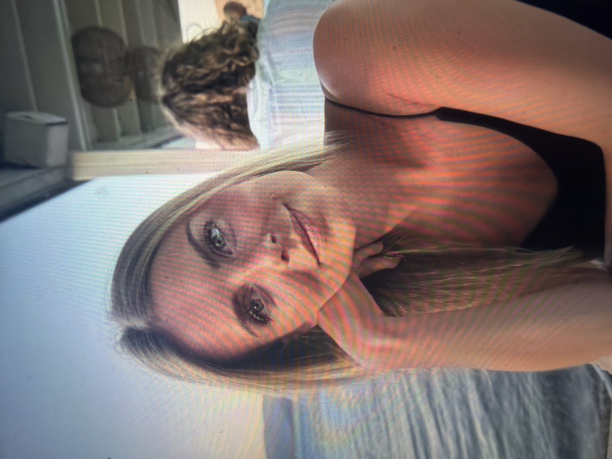The image features a young woman with long, straight blonde hair and light eyes, directly facing the camera with a subtle smile. She is resting her chin on her right hand and is wearing a dark-colored spaghetti strap top. Her skin tones appear warm, possibly due to golden hour lighting or image processing.
To her right and slightly behind her, the back of a second individual's head with curly brown hair is visible. This person is wearing a light-colored, collared shirt and is facing away from the camera.
The background shows an outdoor setting, likely a covered patio or deck, with wooden structural elements including posts and overhead beams. Dark, circular objects, possibly light fixtures, hang from the ceiling. Beyond the immediate structure, a large body of water, resembling a lake or sea, extends to a distant landmass (hills or mountains) under a warm-toned sky, suggesting late afternoon or early evening.
The image itself exhibits clear vertical scan lines and pixelation artifacts, indicating it is a photograph taken of a digital screen rather than a direct photographic capture.
There are no discernible visual elements within the image to confirm a specific location in Shumen, Bulgaria.