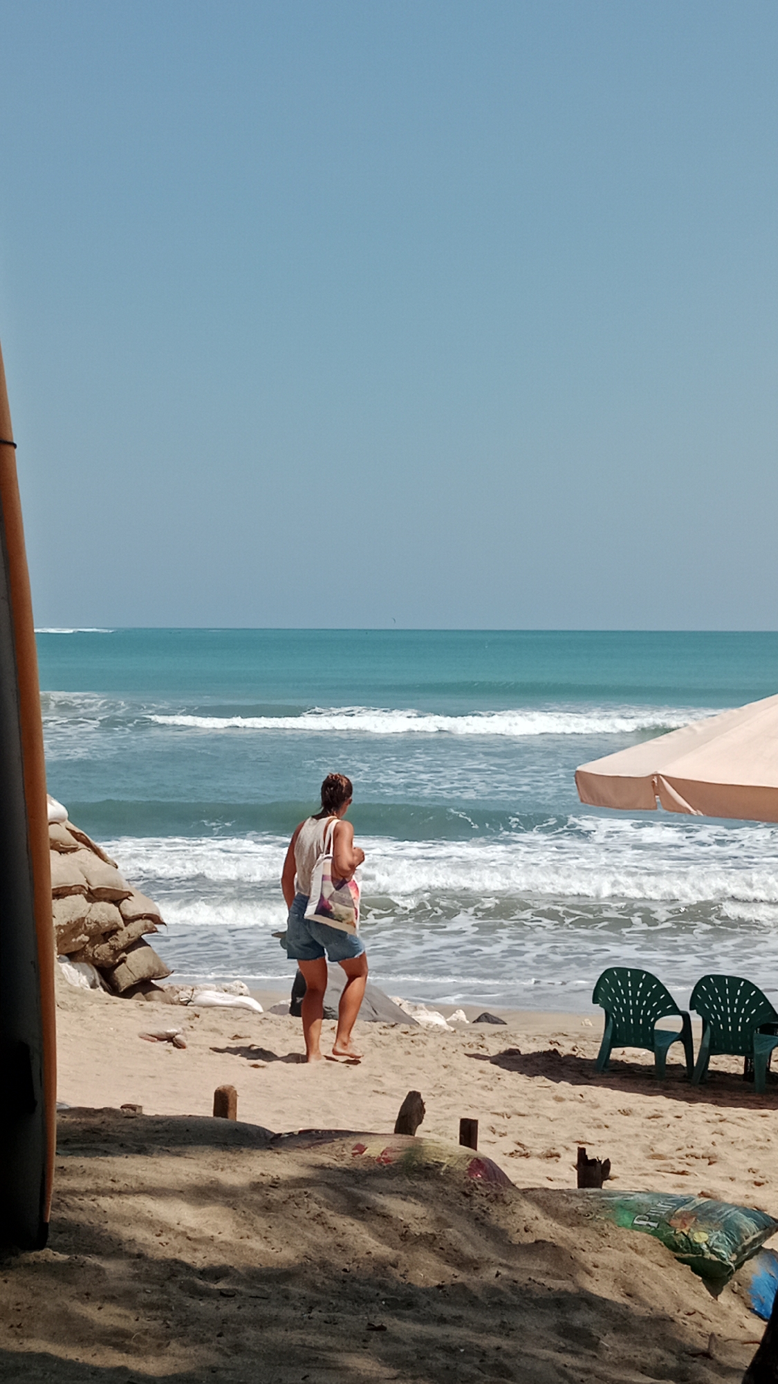 A woman stands on a sandy beach, facing the ocean. She is wearing a light-colored tank top and denim shorts, and she carries a tote bag over her shoulder. In front of her, the turquoise ocean waves crash onto the shore. To her right, a beige umbrella provides shade over two green plastic chairs. To her left, a stack of sandbags is piled on the beach. The sky above is clear and pale blue. The scene appears to be a quiet day at the beach, possibly in a tropical or coastal location.