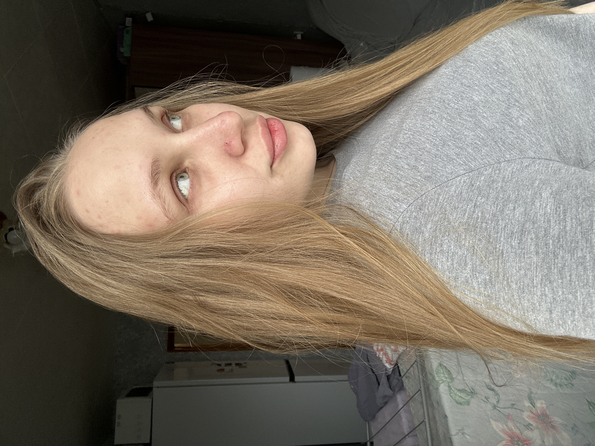 A young woman with long, straight, light blonde or light brown hair is the central subject of this image, captured in what appears to be an indoor setting. She is positioned with her head tilted back, looking upwards and slightly to her right, suggesting she is reclining or lying down. Her fair skin shows a few small red blemishes on her forehead and nose. Her eyes are light-colored, possibly green or blue-green, and her lips are naturally colored and slightly parted, contributing to a neutral, perhaps pensive or calm, expression. She is wearing a plain grey, long-sleeved top.
The background is softly blurred and dimly lit, indicating an indoor environment. On the right side of the frame, a dark, possibly wooden, piece of furniture or shelving is visible. Towards the bottom left, a floral patterned fabric, likely a bedsheet or blanket, adorned with pink flowers and green leaves, suggests a bedroom or living space. Other elements in the background are indistinct due to the low light and shallow depth of field. The lighting is soft and ambient, without strong shadows, indicating an indoor scene during daytime. There is no visible text in the image, and no specific visual cues within the photo itself point to the given location of Chaikovsky, Russia. The overall mood is quiet and still.