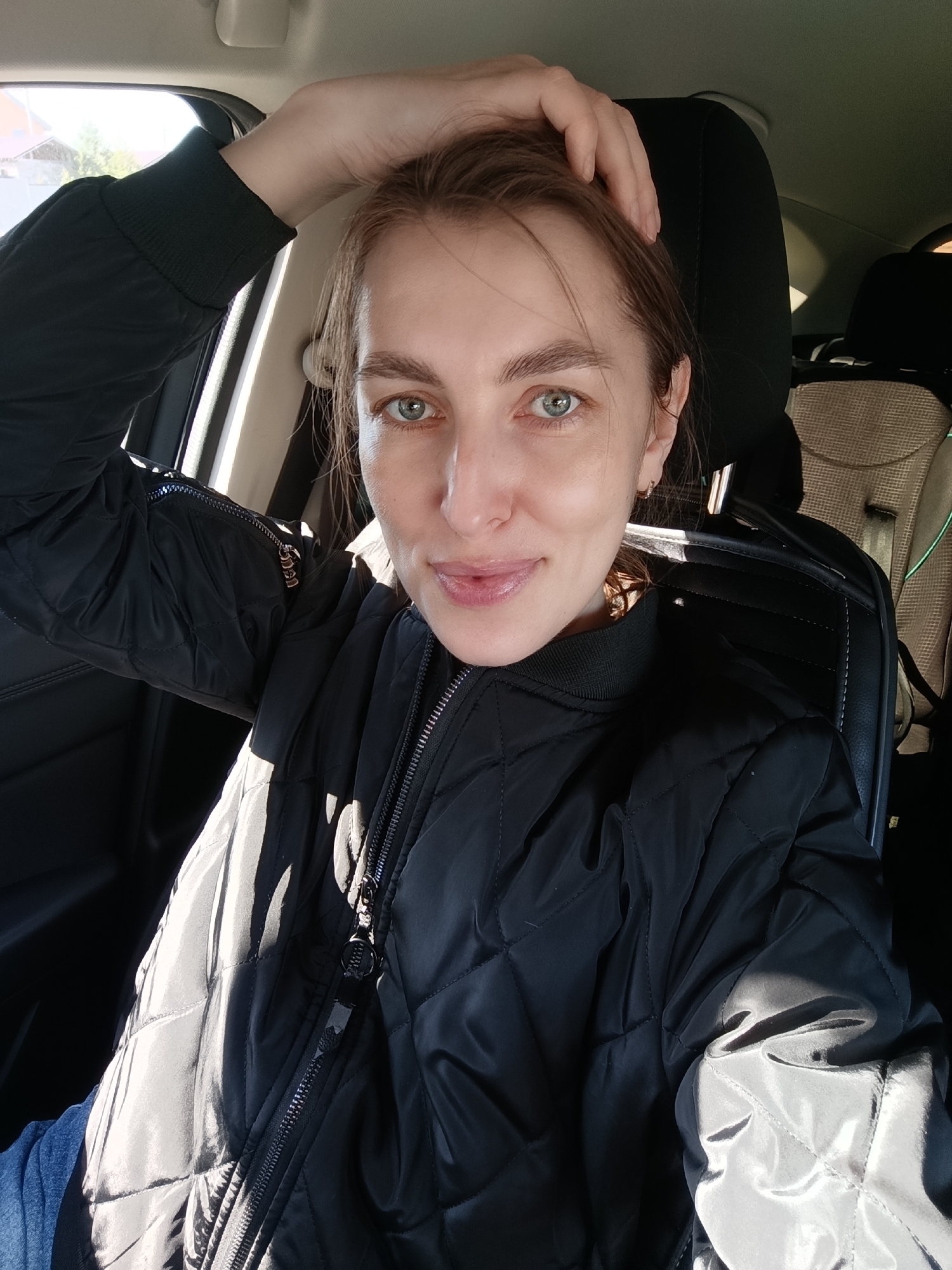 A fair-skinned woman with light brown hair pulled back from her face is sitting in the driver's seat of a car. She has blue-green eyes and appears to be wearing minimal makeup. Her right hand is resting on the top of her head.
She's wearing a black, quilted bomber jacket with a zipper that is partially unzipped. Sunlight is illuminating the left side of her jacket and face, creating shadows.
The car's interior is visible, including the black leather seats and the headrest behind her. A tan-colored car seat is visible in the back.
Based on the provided information, the location is Chaikovsky, Russia, although no specific geographical details are visible in the image to confirm this.