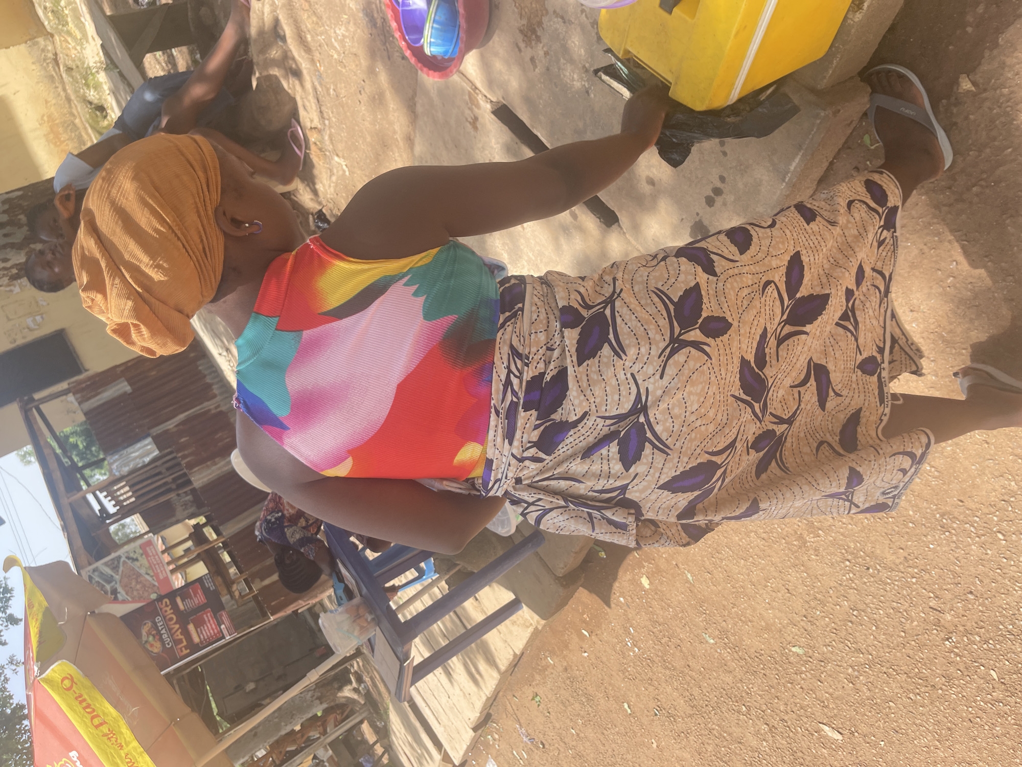 The image shows a street scene in Rafin Par, Nigeria. A woman in a brightly colored top and patterned skirt is standing and reaching into a yellow container. She is wearing an orange headscarf and flip-flops. In the background, there is a stall with an umbrella advertising "Curated Flavors." Other people are visible in the background, some sitting and others standing. The ground is dirt and gravel, and there are several plastic tables and chairs. A red bowl with other bowls inside is also visible.