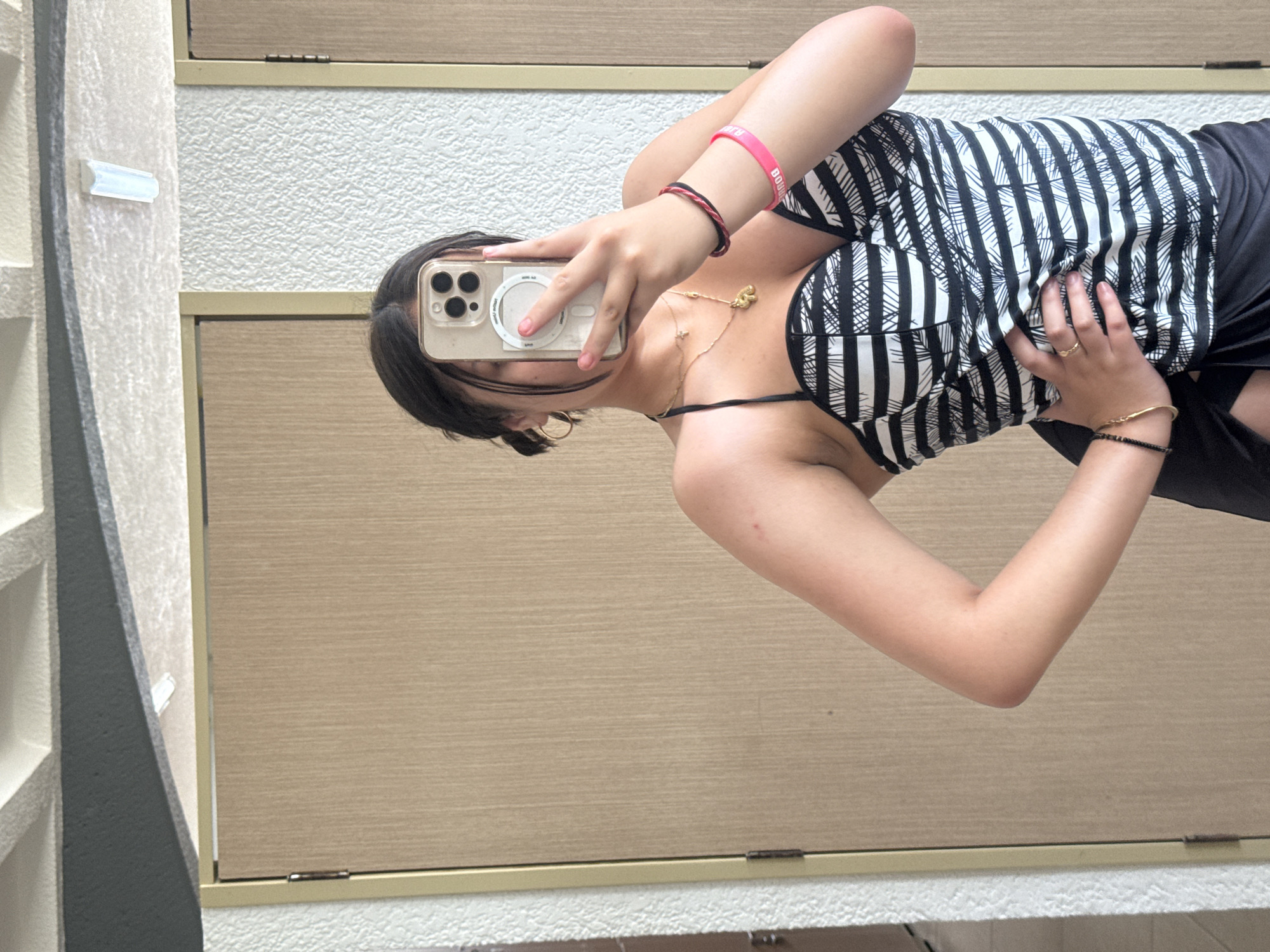 A mirrored image shows a young person with dark hair wearing a striped black and white top and black bottoms. They are holding a phone up to their face, obscuring it, and taking a selfie. The background includes light-colored, textured walls and wood-paneled surfaces. The location is Unknown, Unknown.