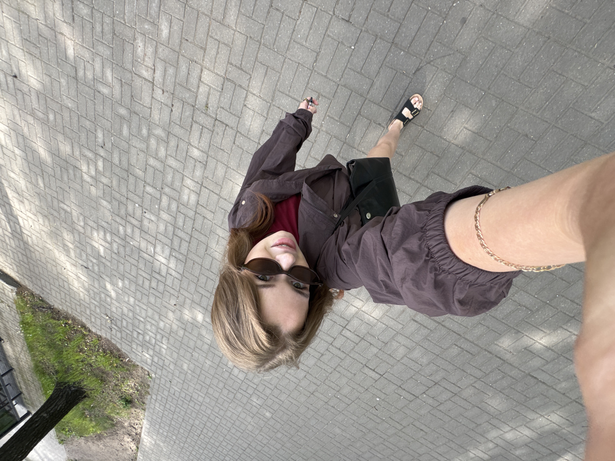 A young woman with shoulder-length brown hair and sunglasses is captured from a high-angle perspective, possibly a selfie taken while walking. She is wearing a dark brown jacket over a burgundy top, a black skirt, and black sandals. A black purse is slung over her shoulder. She is walking on a paved sidewalk in Chaikovsky, Russia, with a tree and green grass visible to the left. The sunlight casts shadows on the ground, indicating it is daytime.