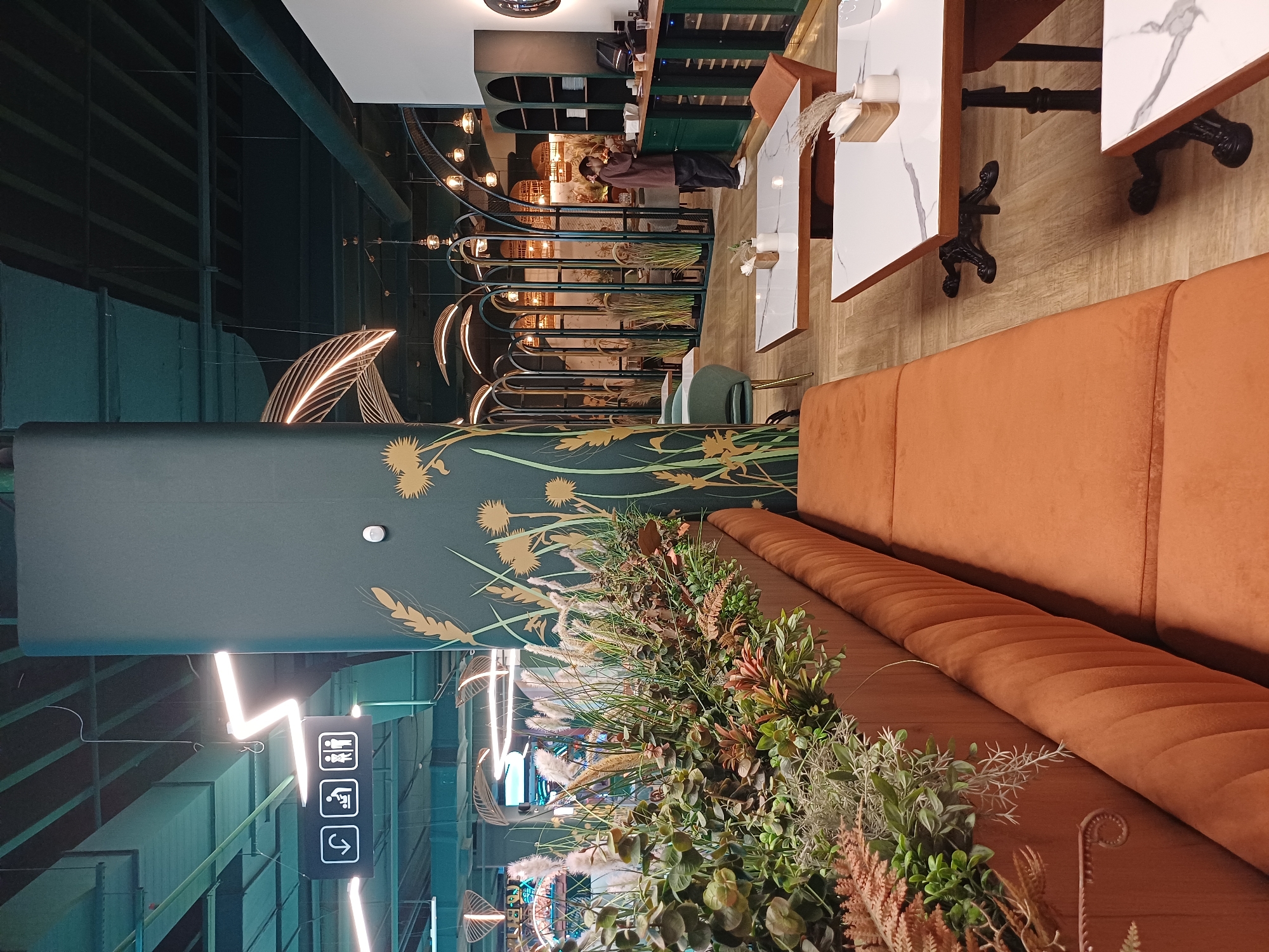 A dimly lit, modern restaurant interior features a long, plush orange banquette seating area along the left side of the frame, adjacent to a raised planter filled with artificial greenery. The planter is set against a large, dark green pillar decorated with a golden wheat stalk motif. Beyond the banquette, several tables with white marble tops and ornate black bases are visible, set for dining. In the background, a person in a brown uniform is attending to a counter area with green shelving units and wine coolers. The ceiling is a dark green, exposed industrial structure with hanging lights and ventilation systems. Archways and decorative screens create a sense of partitioned space in the mid-ground, leading to a more distant seating area. Signs indicating restrooms and a changing station are suspended from the ceiling in the upper left portion of the image. The overall ambiance is contemporary and sophisticated, with a touch of naturalistic decor.