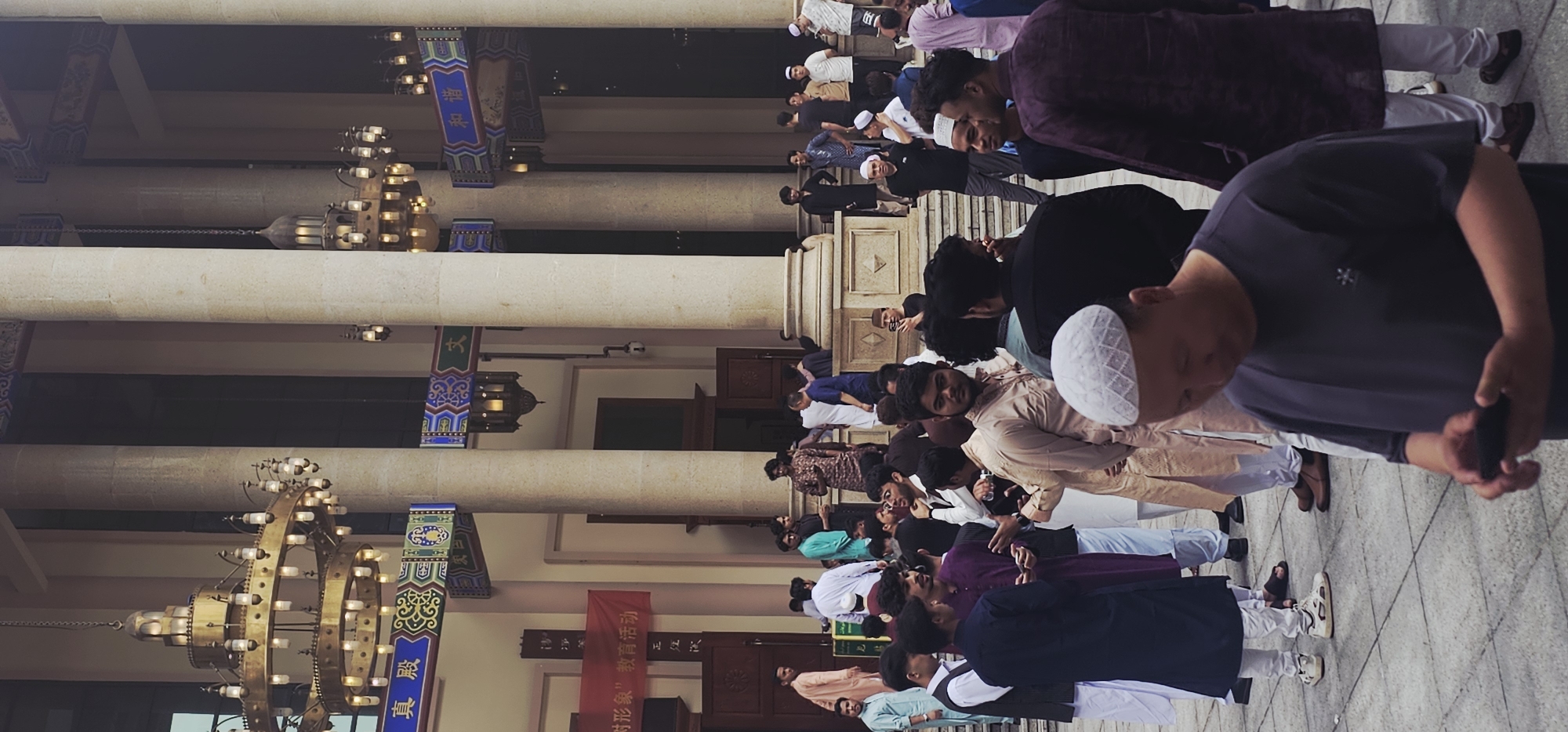 The image shows a group of men praying in what appears to be a mosque or religious building in Jiulongpo District, China. They are lined up in rows, some wearing traditional Muslim attire like kufis (skullcaps). The building features large pillars, ornate chandeliers, and Chinese script on decorative banners, suggesting a blend of architectural styles.