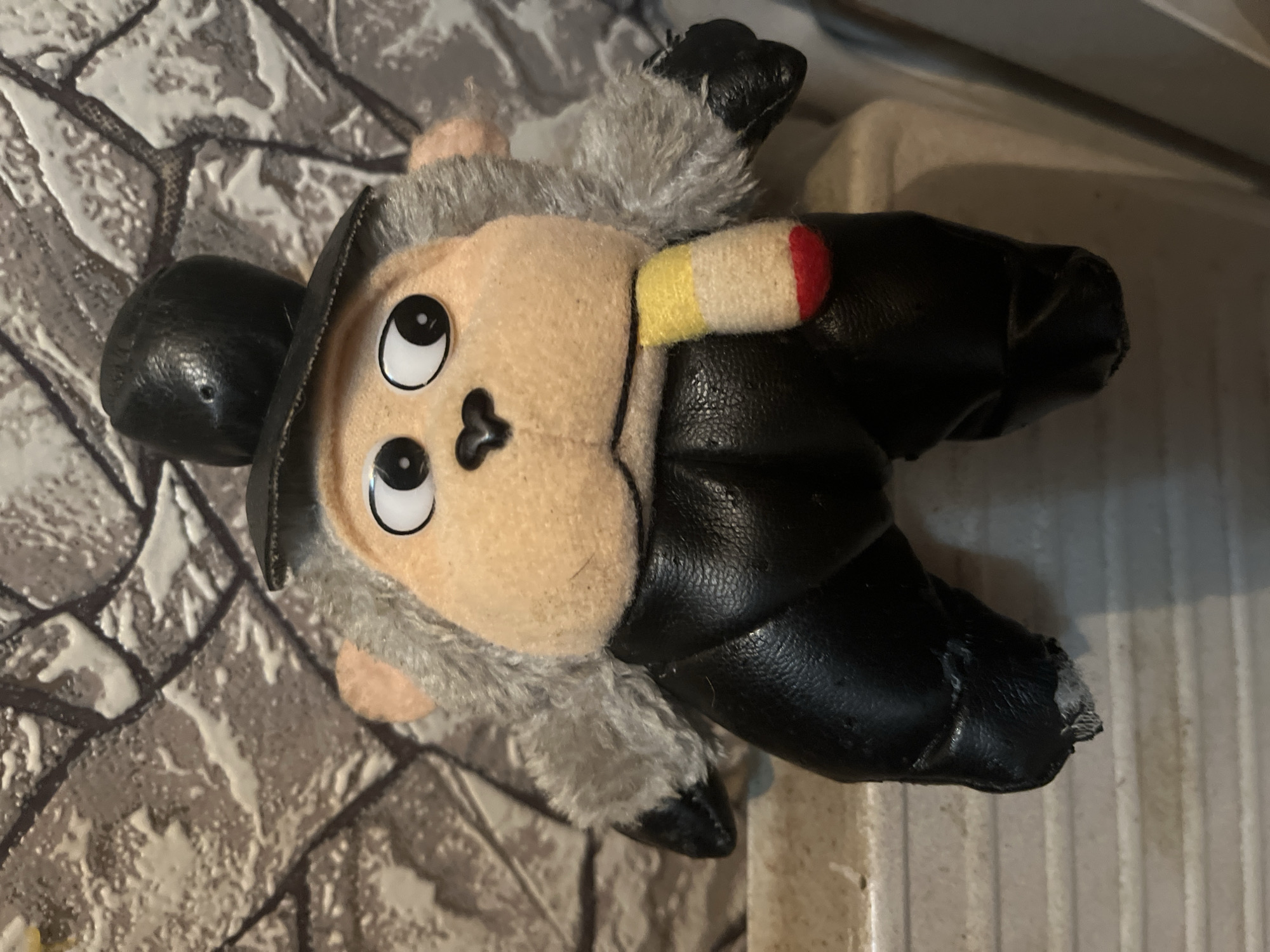 A plush toy monkey wearing a black top hat and black pants sits on a white ledge. The monkey has grey fur around its face, a tan face with large white eyes with black pupils, and a black nose. It is wearing a scarf with yellow, white, and red stripes. The monkey's left leg appears damaged, with the black material torn and frayed. The background is a textured grey wall that resembles stone. The scene does not contain any identifiable landmarks or features suggesting it is located in Minsk, Belarus.