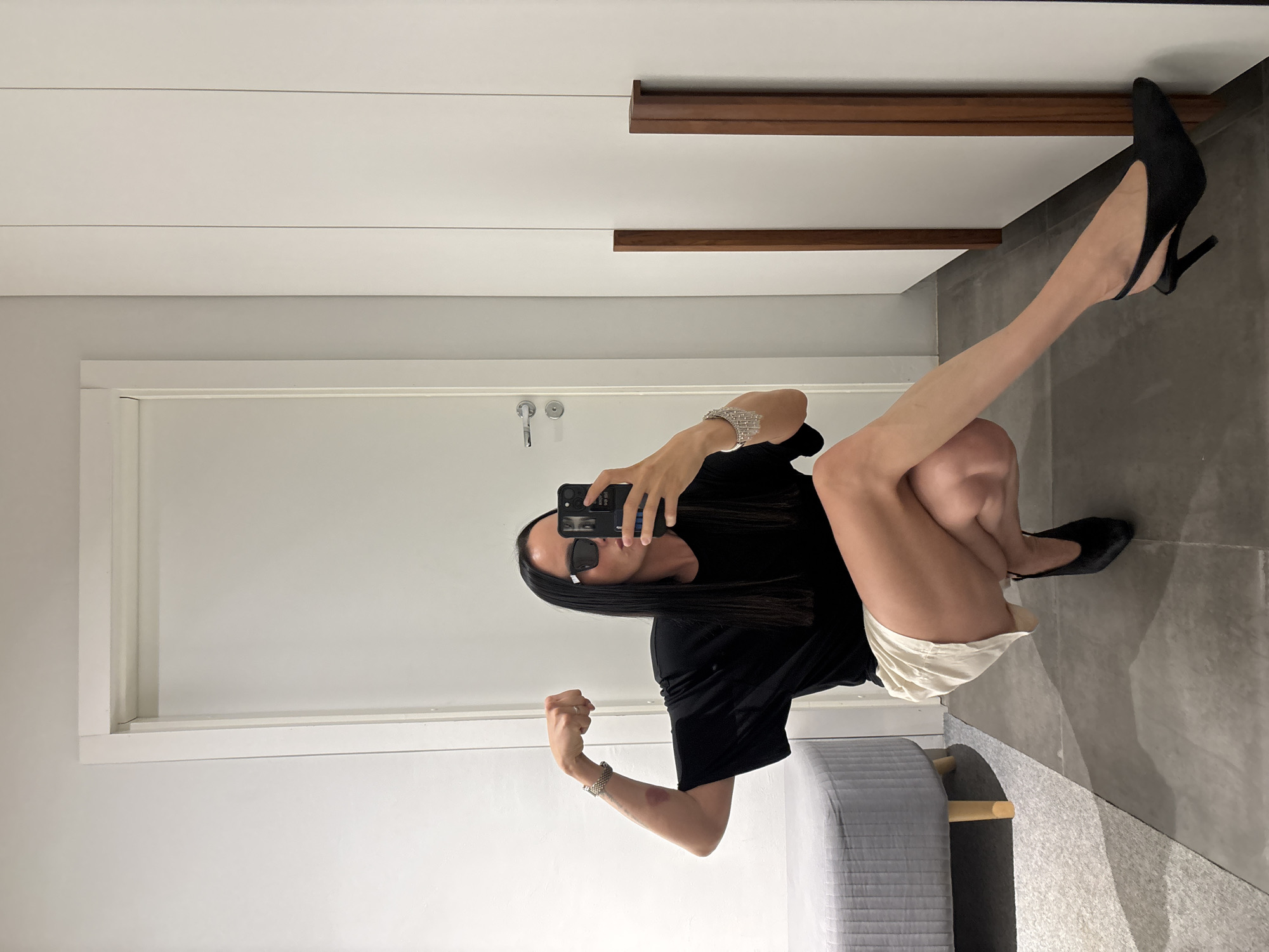 A woman is squatting and posing for a photo in an indoor setting. She is wearing a black t-shirt, white shorts, and black heels. She is holding a smartphone in her hand and looking at the camera. Her right arm is raised with her fist clenched. A lipstick mark is visible on her right forearm. She is also wearing sunglasses. The setting appears to be a modern interior, with a white door and wall in the background and a gray chair. There is a cabinet or closet with wooden handles above her. The floor is tiled with gray stones. The location is unknown, and there is no information about the city of Chaikovsky, Russia, present in the picture.